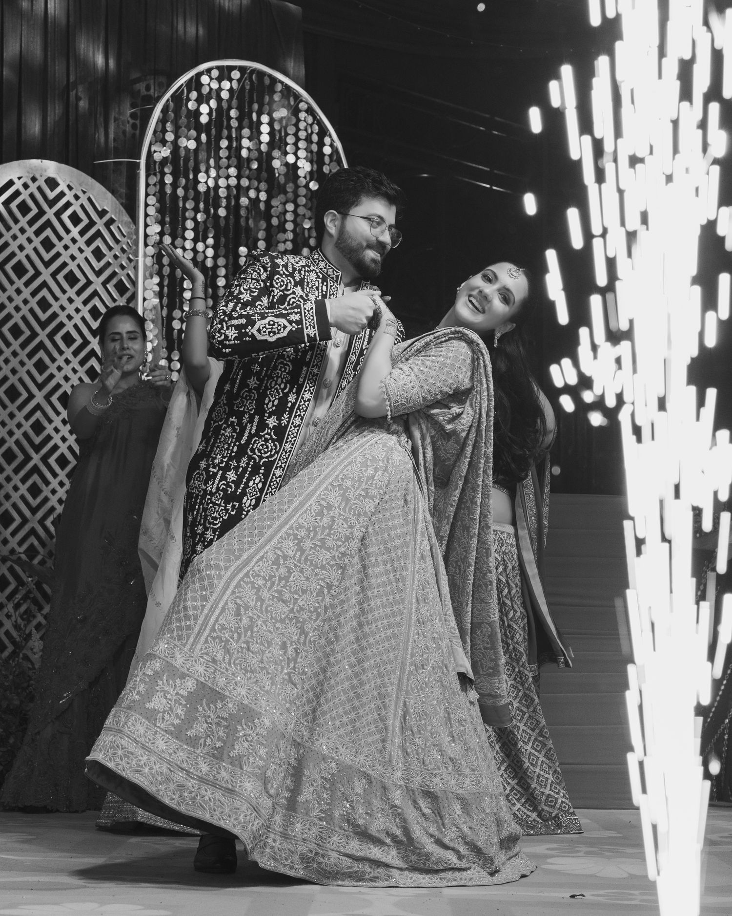 Couple dancing joyfully in traditional Indian attire at a wedding celebration, black and white photo.