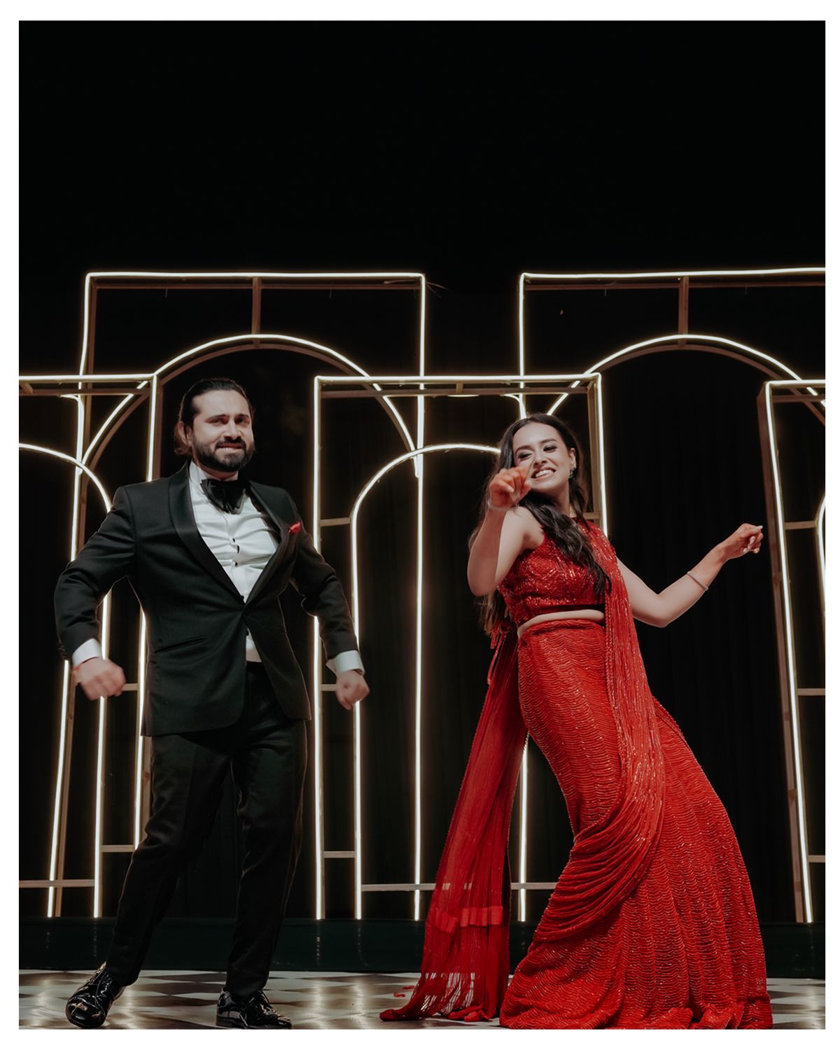 A couple performing on stage with golden arches, woman in red gown and man in black suit striking dramatic poses.
