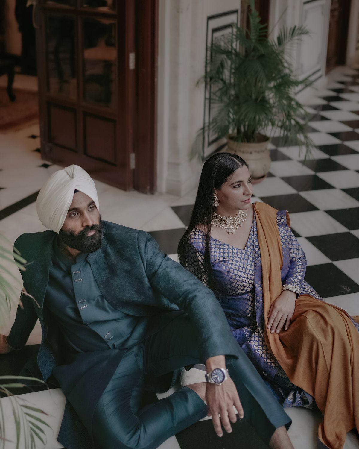 A couple in traditional Indian formal wear relaxes on seating in an elegant interior with checkerboard flooring.