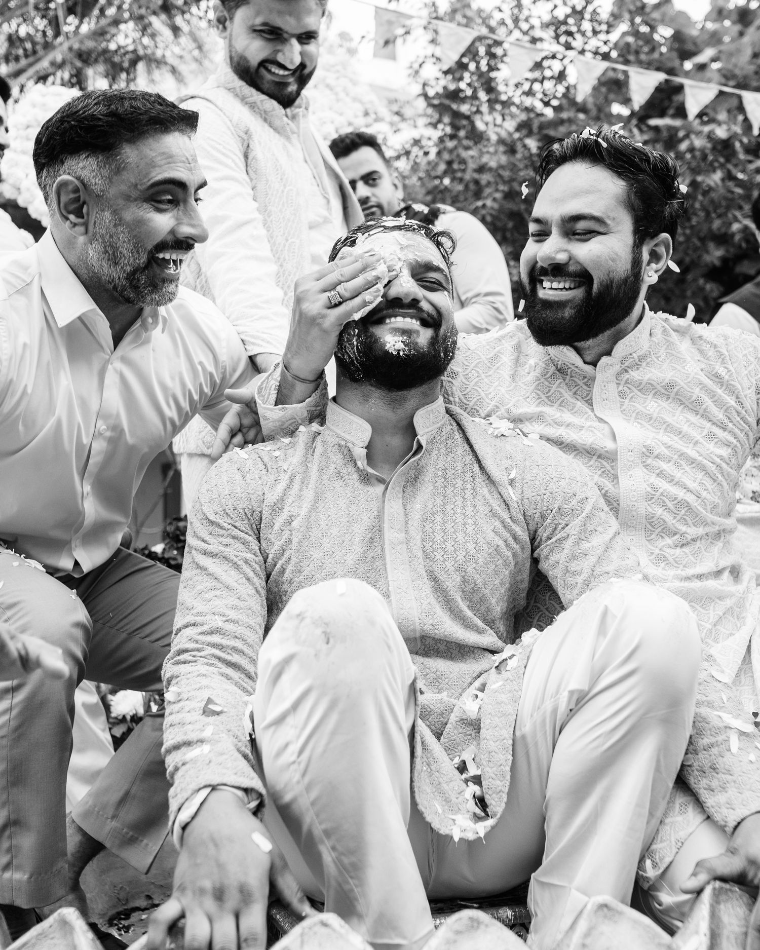 Black and white photo of men laughing during a joyful Indian wedding celebration ceremony outdoors.