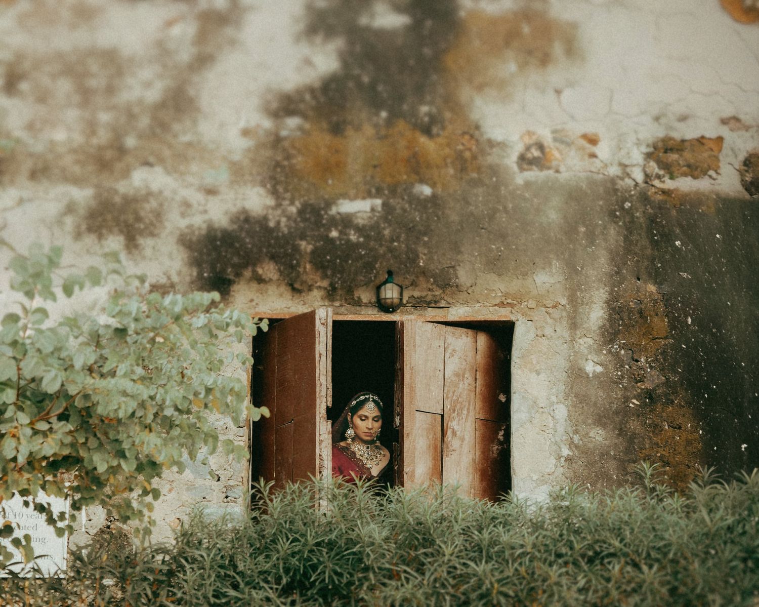 A woman in a red sari stands in a rustic doorway surrounded by lush greenery and a weathered stone wall.