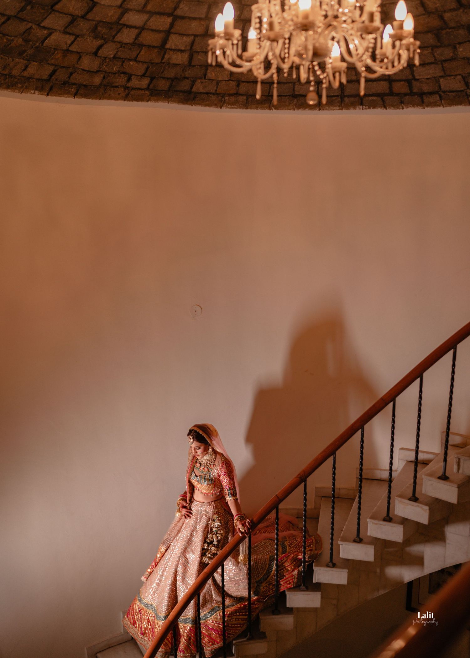 Artistic silhouette of a bride in traditional attire descending a staircase under a crystal chandelier.