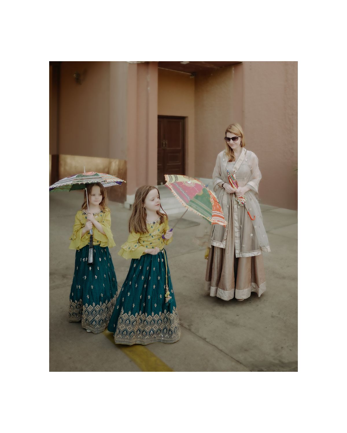 Three people in traditional Indian attire holding parasols pose together outdoors.