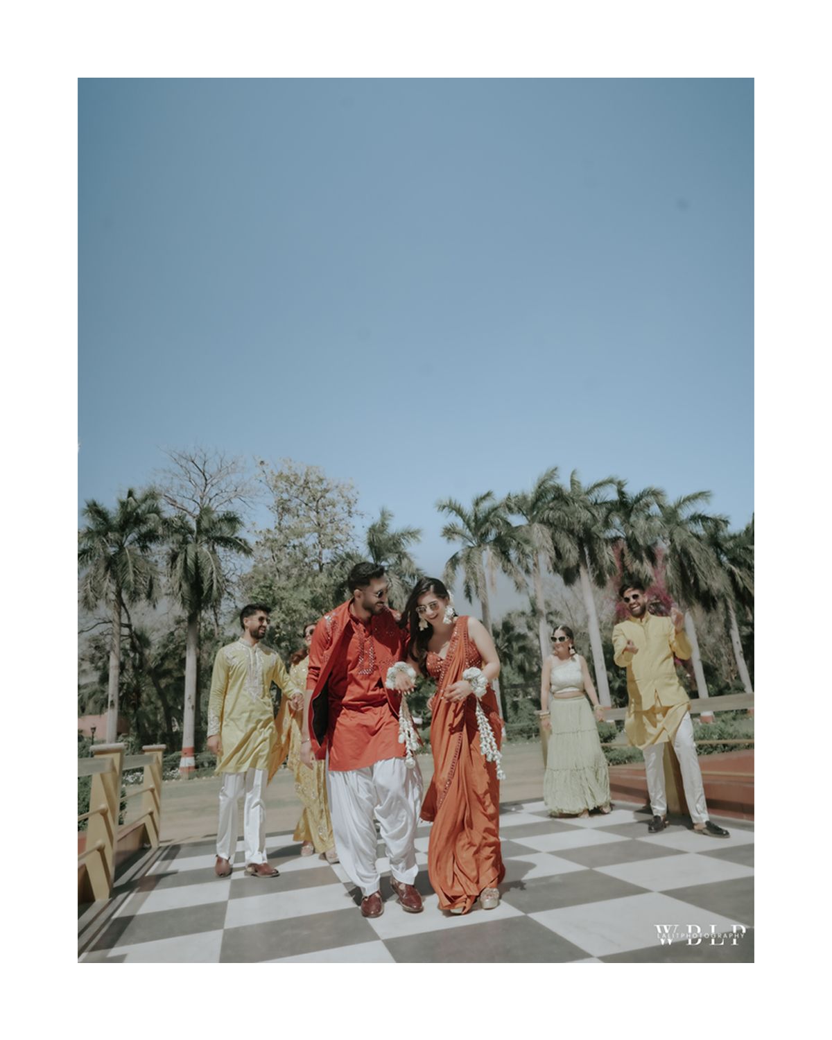 Group celebrating on outdoor checkered patio with palm trees and traditional Indian formal wear.