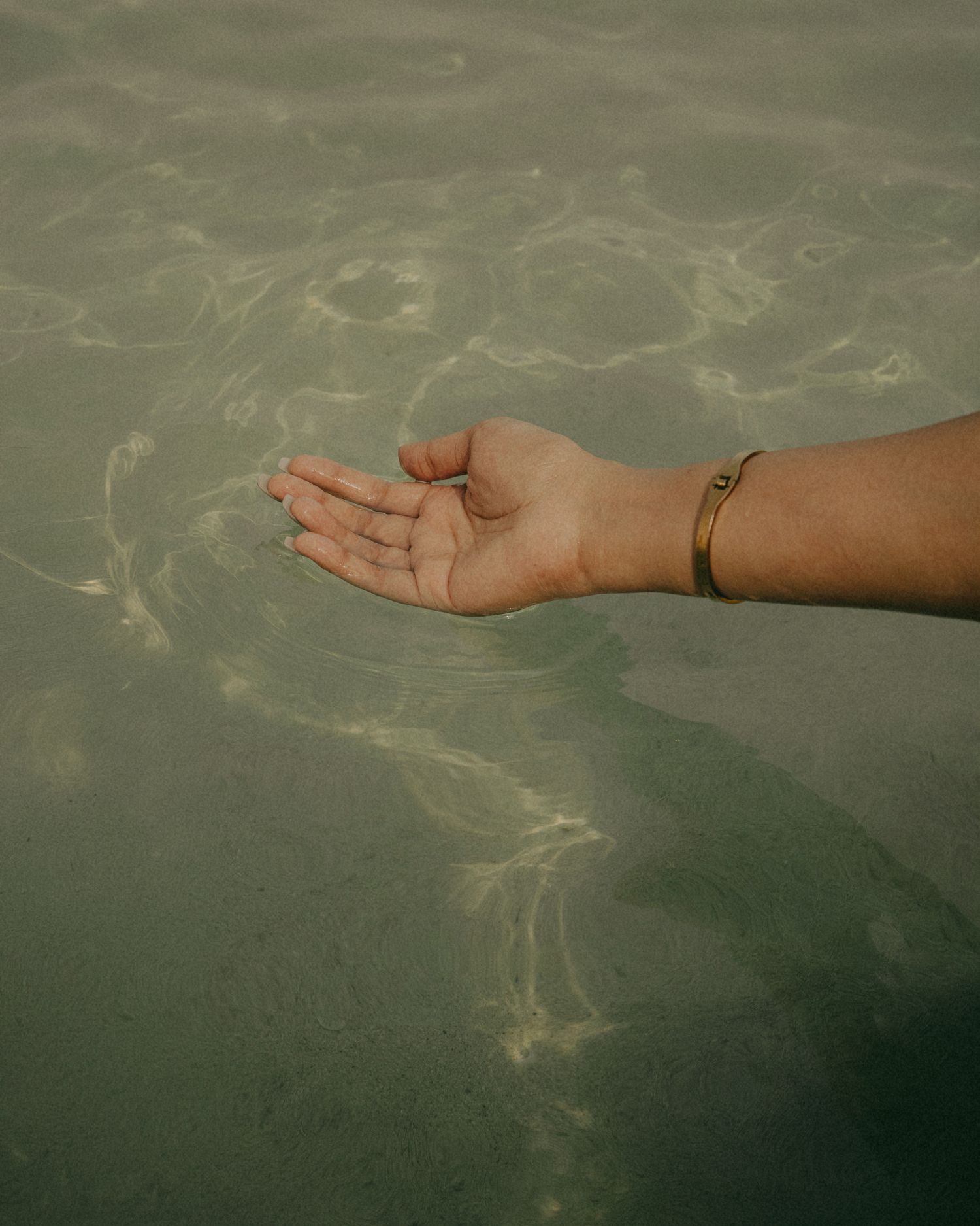 A hand with a gold bracelet gracefully extended over calm, sunlit shallow water with rippling reflections.