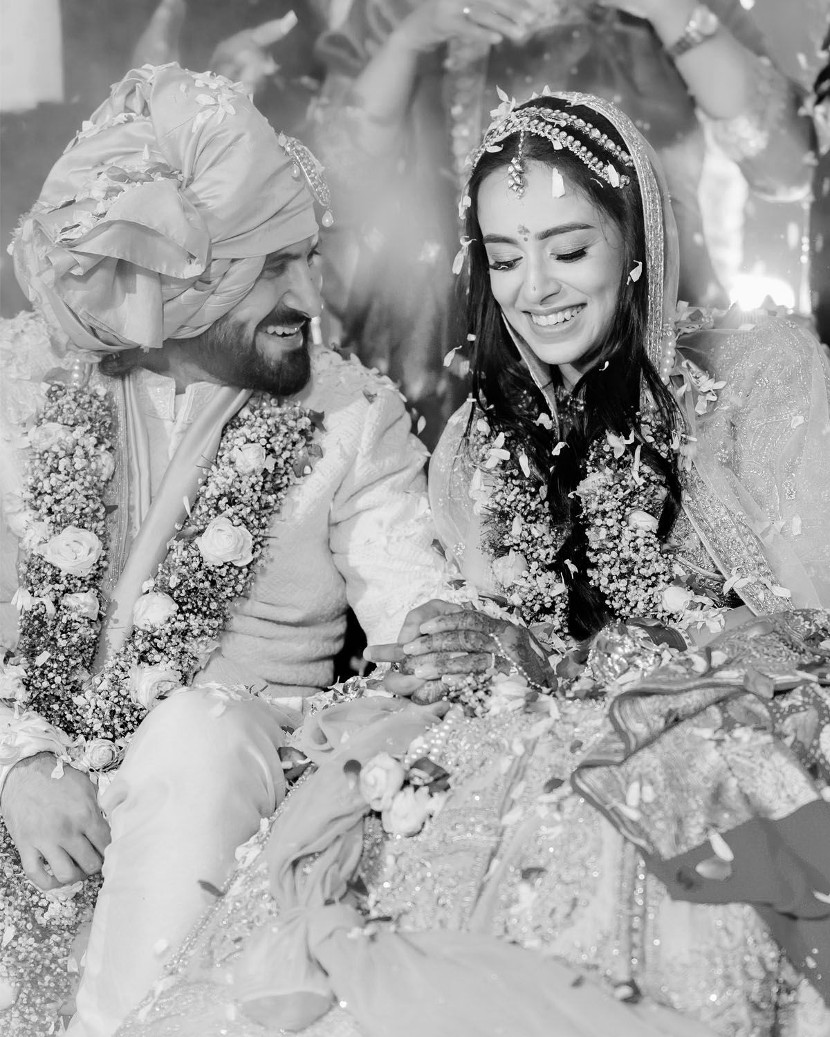 A joyful couple in traditional Indian wedding attire share a candid black and white moment amid falling flower petals.
