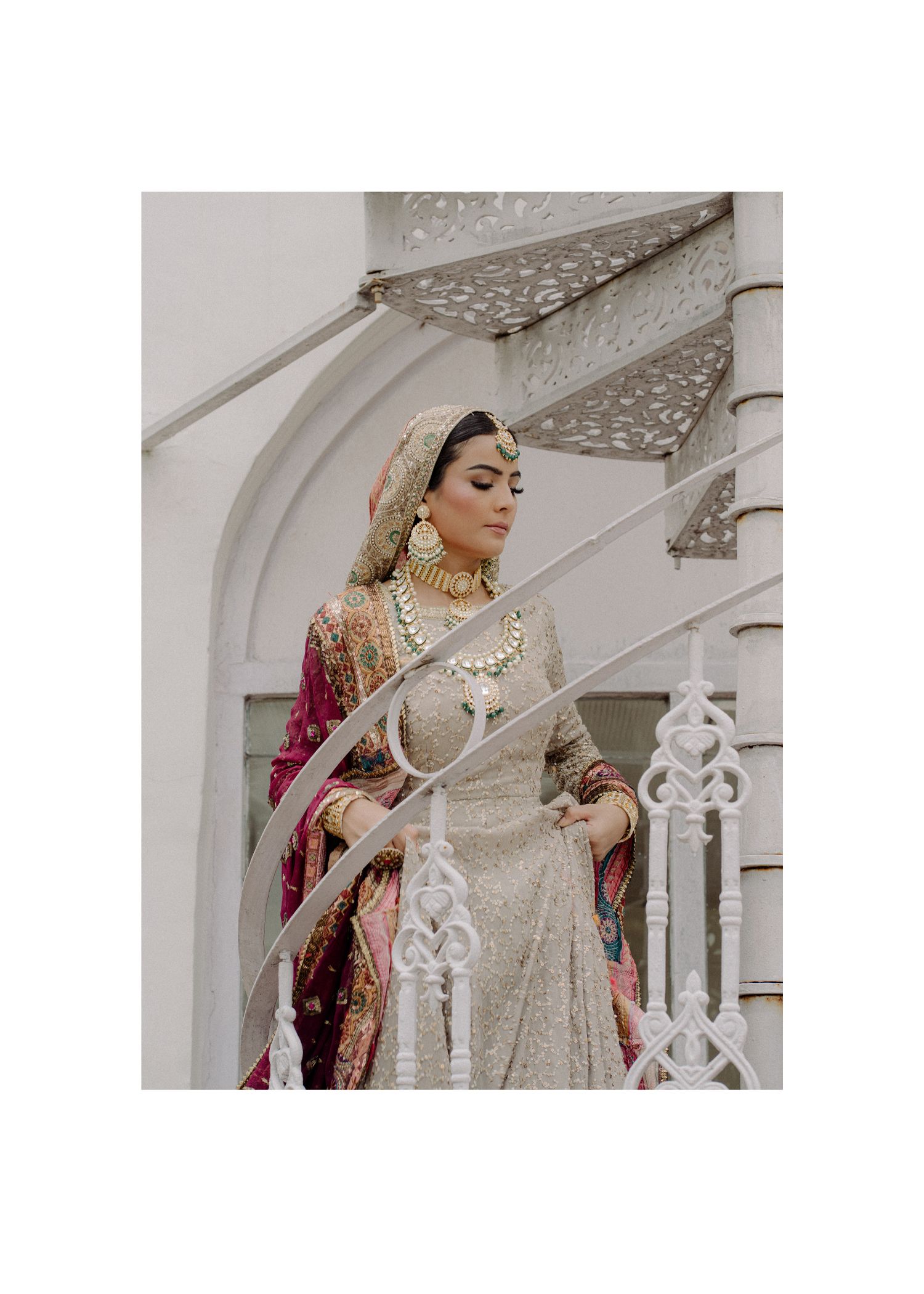 A bride in an elegant beige lehenga and red dupatta poses against a white architectural backdrop with ornate railings.