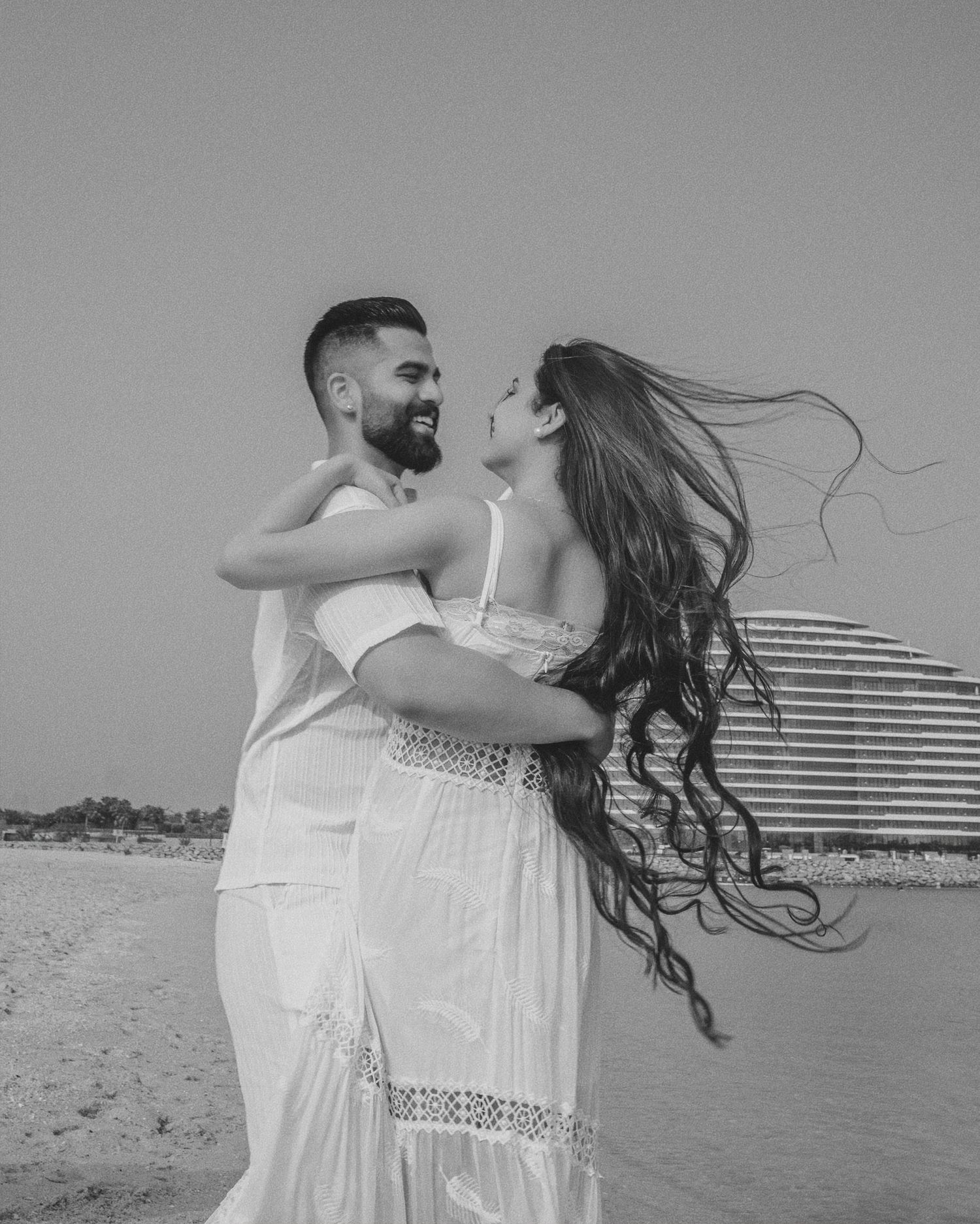 Black and white photo of a couple embracing on a beach, woman's long hair blowing in the wind.