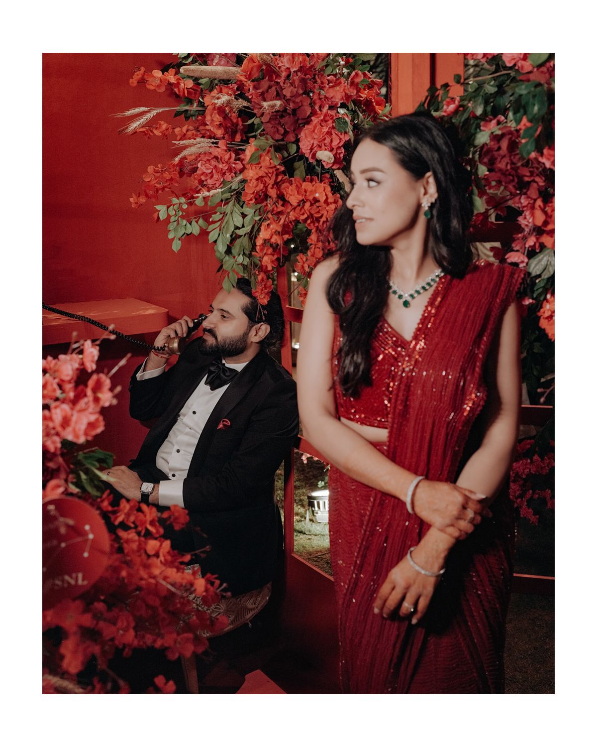 A couple in formal attire pose against a backdrop of vibrant red flowers and wall, creating a dramatic romantic setting.