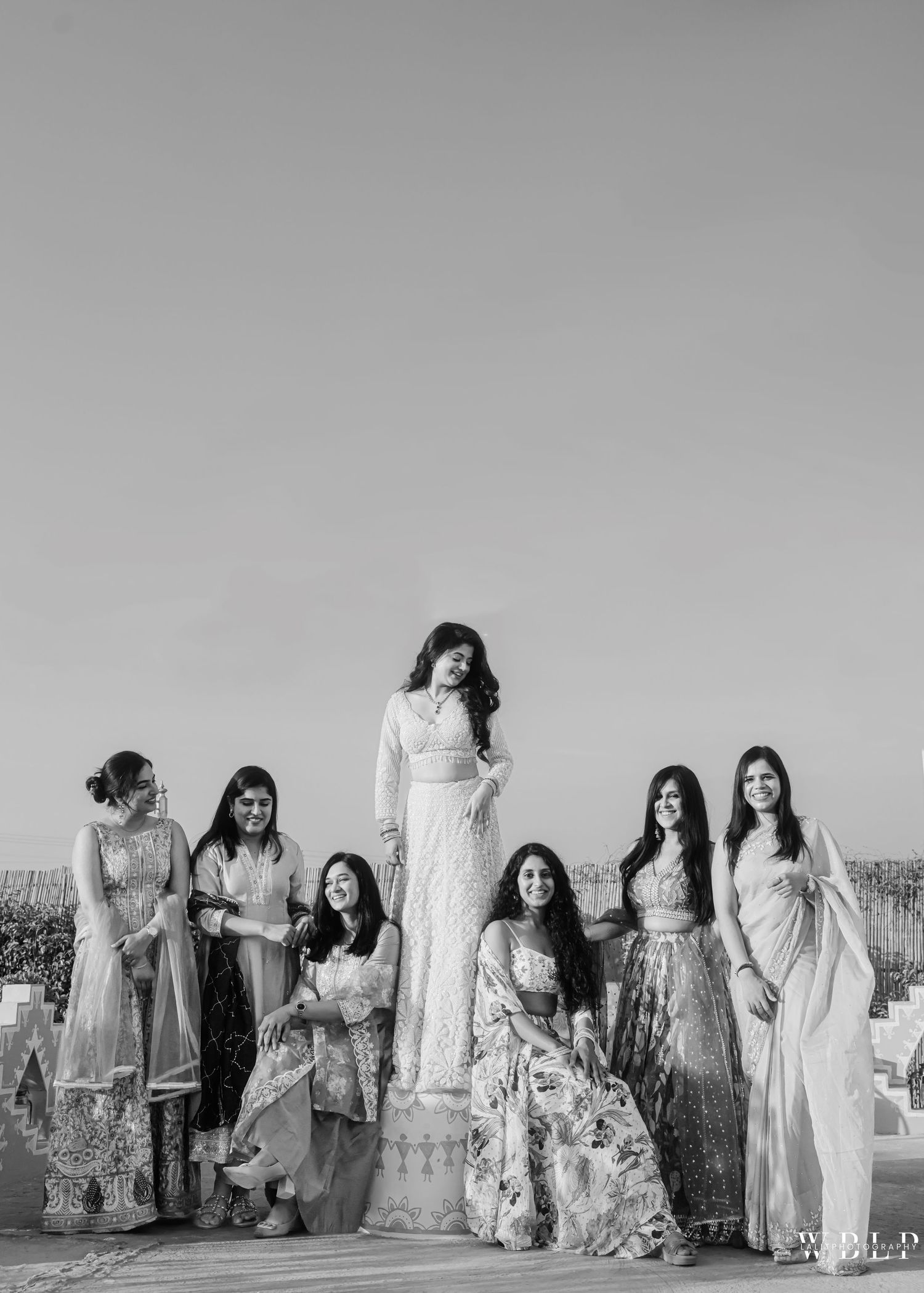 Black and white photo of a bride in white lehenga posing with six bridesmaids outdoors at an Indian wedding.