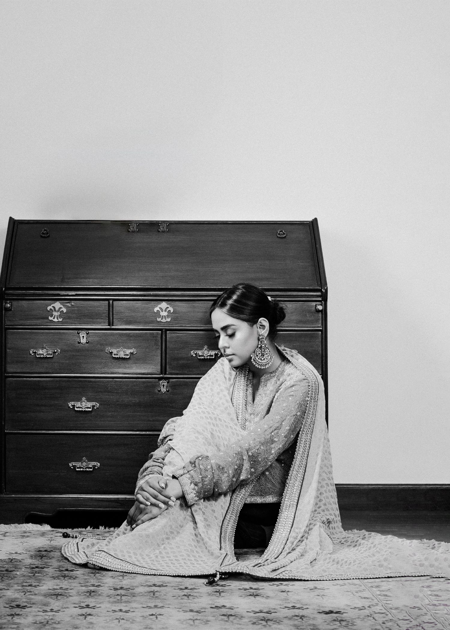 Black and white vintage photograph showing elegant figure reclining on carpet beside wooden dresser in contemplative pose.
