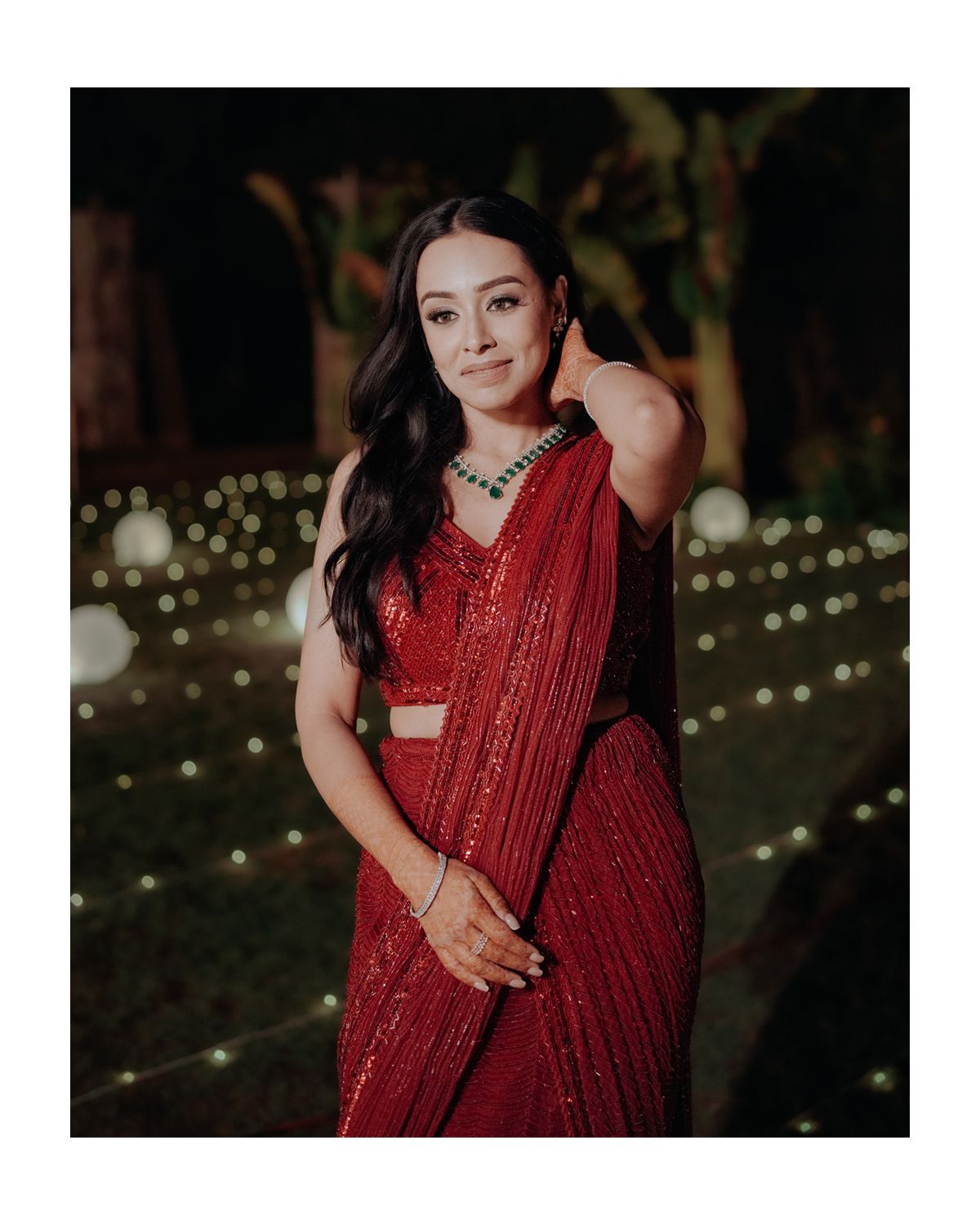 A person in a sparkly red formal sari poses elegantly against a backdrop of outdoor string lights in an evening setting.