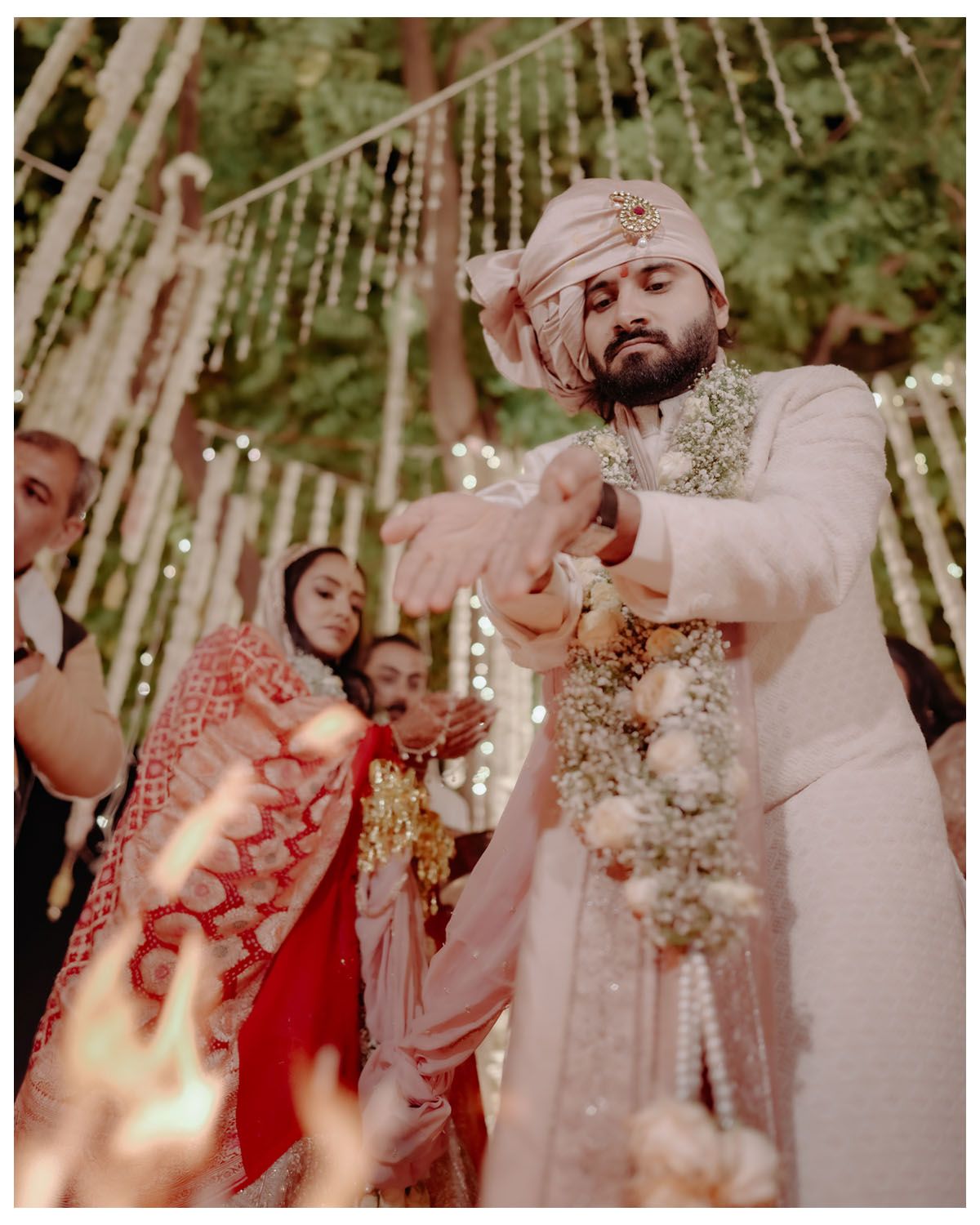 A couple dressed in elegant Indian wedding attire celebrates during a ceremony under hanging floral decorations.