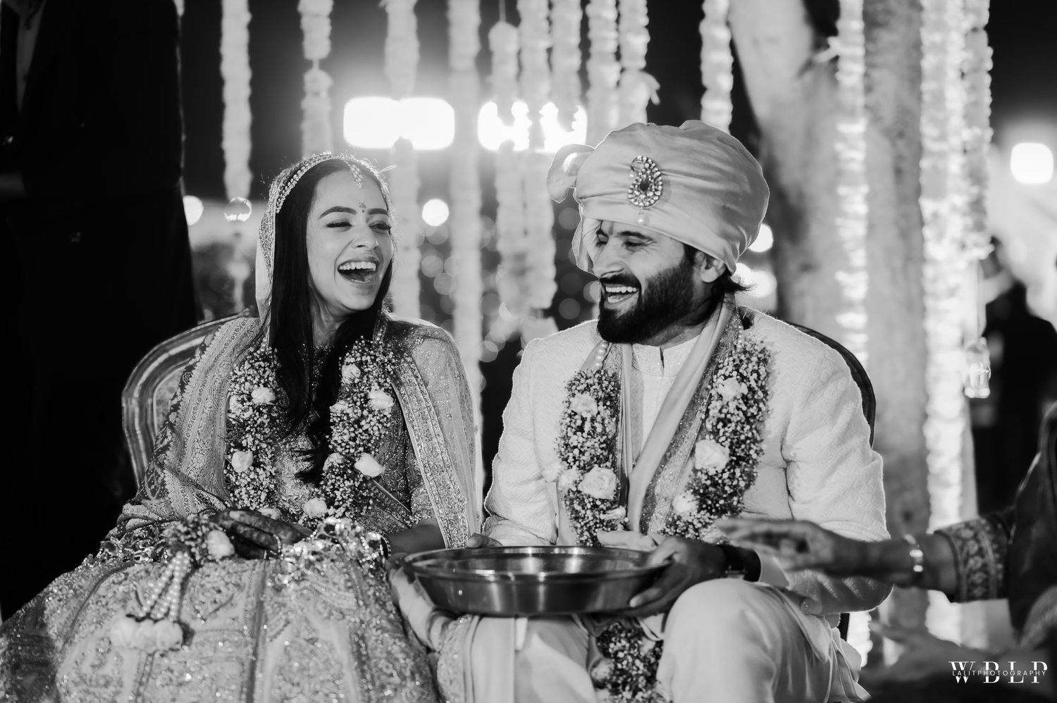 A couple in traditional Indian wedding attire share a joyful moment during their ceremony with glowing lights in the background.