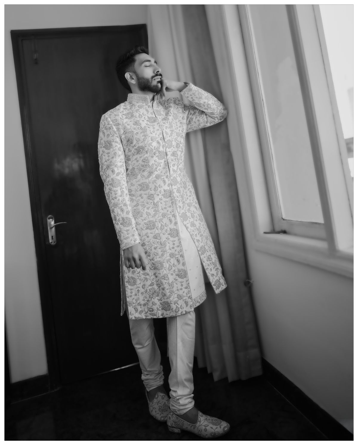 A man in embroidered white traditional Indian formal wear poses against a wall.