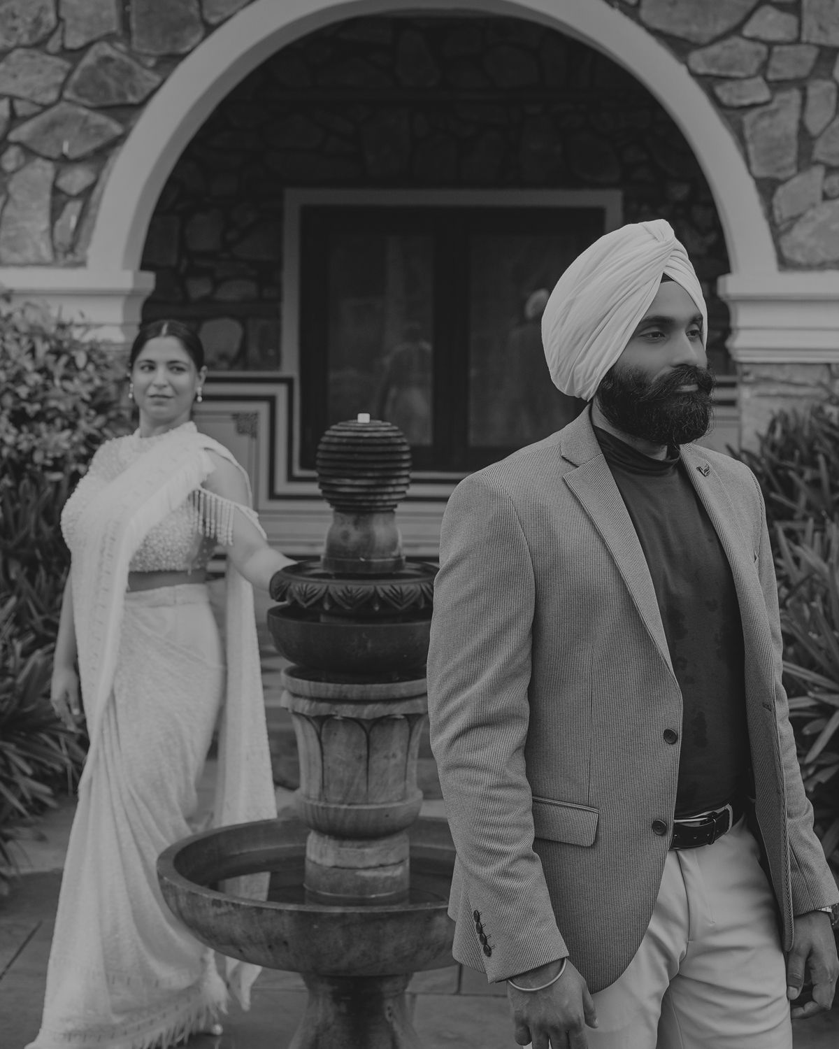Black and white portrait of a couple standing by a fountain outside a stone building.