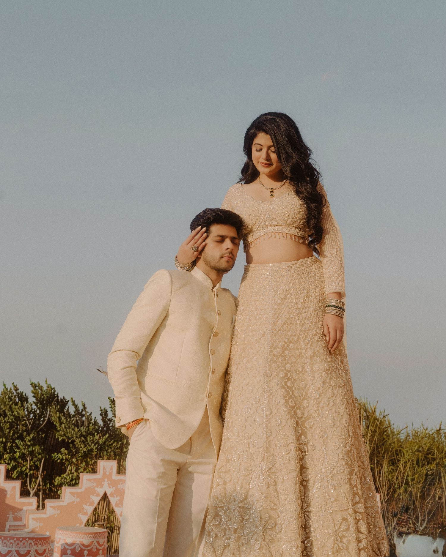 Couple in elegant cream Indian wedding attire posing outdoors, woman standing elevated beside seated man.