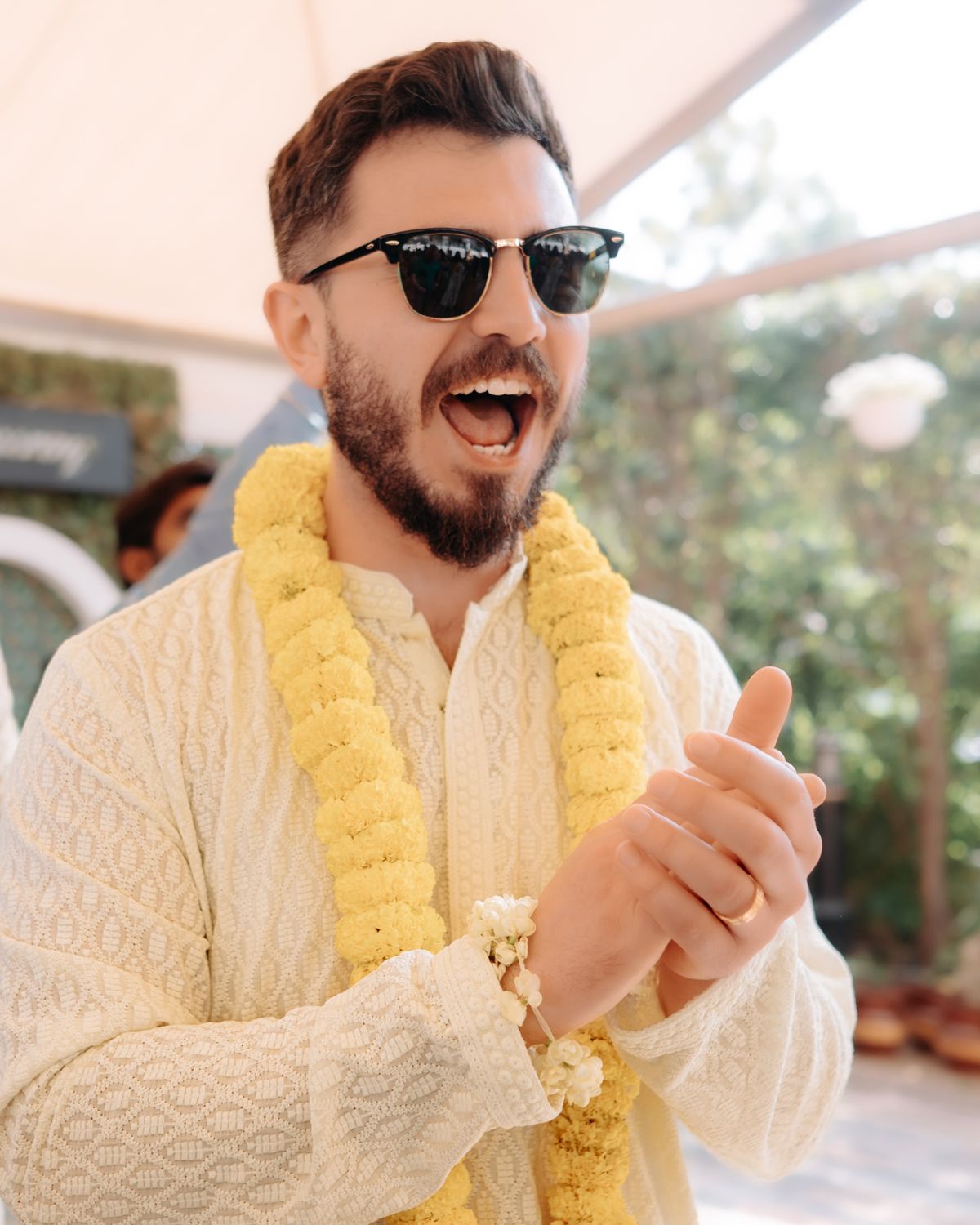 Person wearing sunglasses and white outfit with yellow garland laughs joyfully at outdoor celebration.