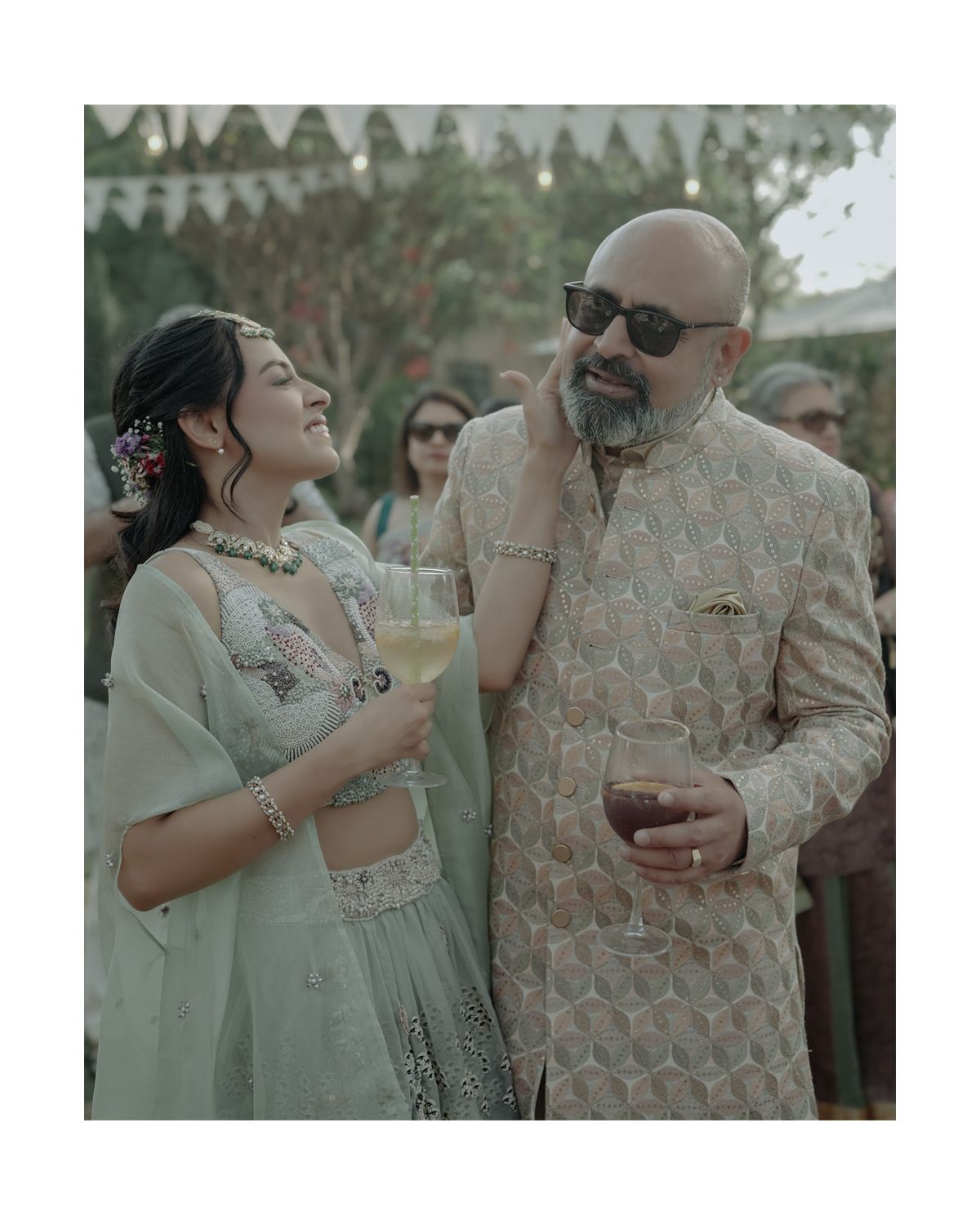 Two guests in elegant Indian attire enjoying drinks at an outdoor celebration with festive decorations.