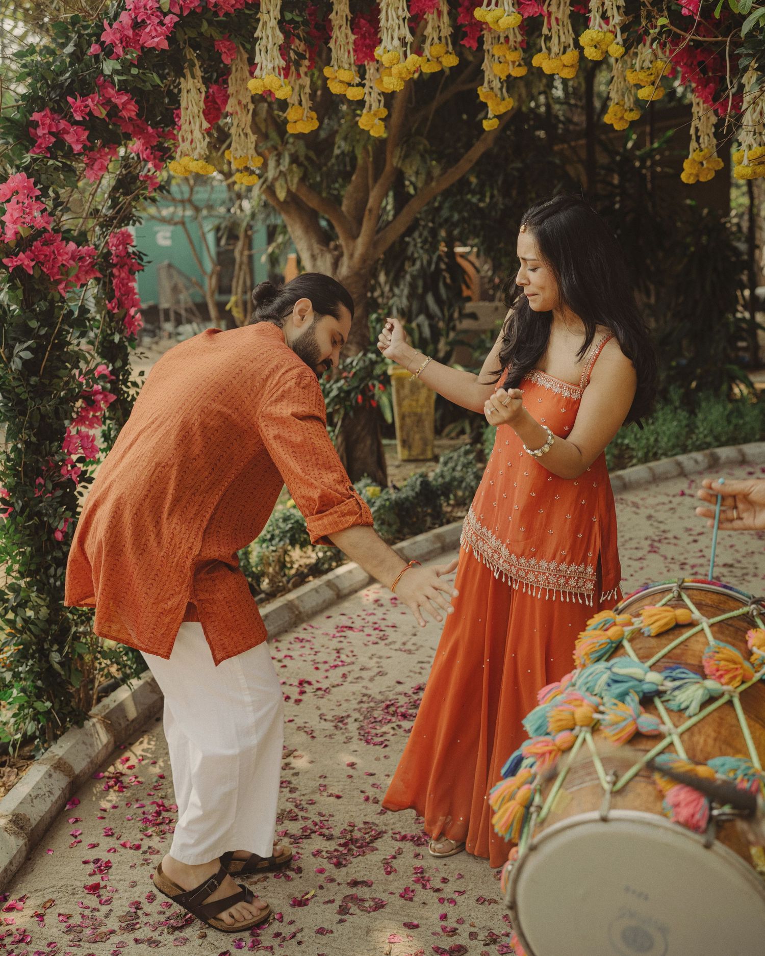 A couple in matching orange outfits celebrates during a traditional Indian ceremony with floral decorations and drums.