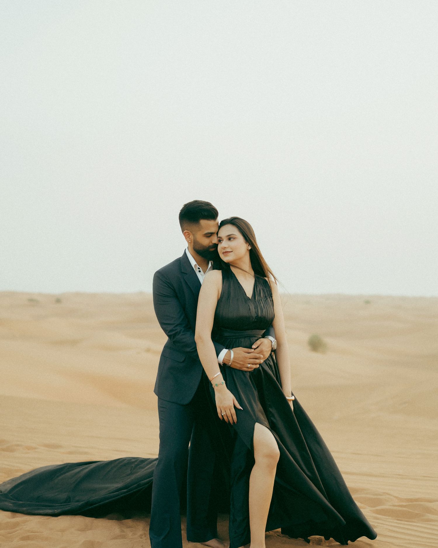 Couple in black attire poses romantically in a desert landscape with sand dunes and overcast sky.