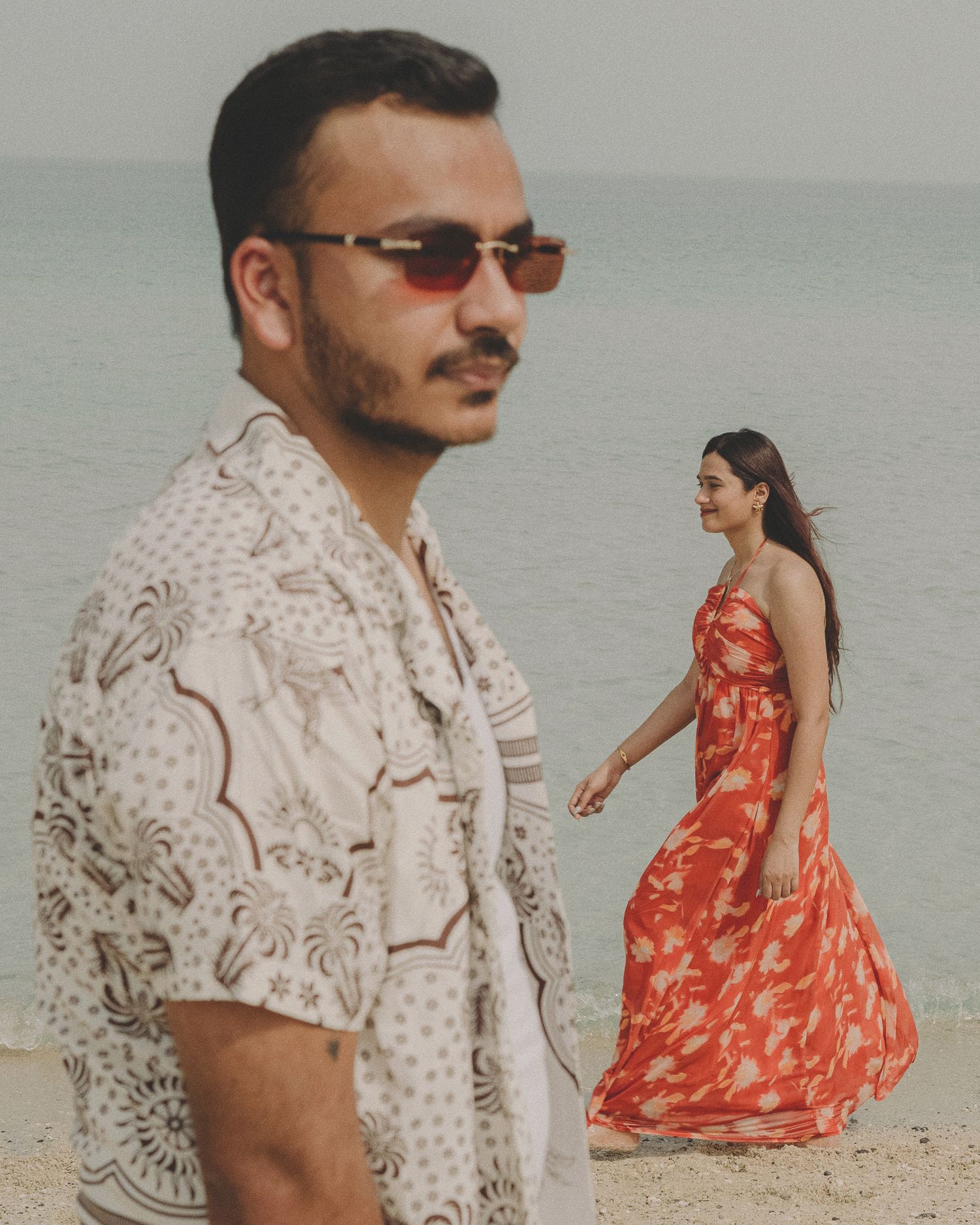 A man in a printed shirt and sunglasses stands on a beach while a woman in a red floral dress walks behind him.