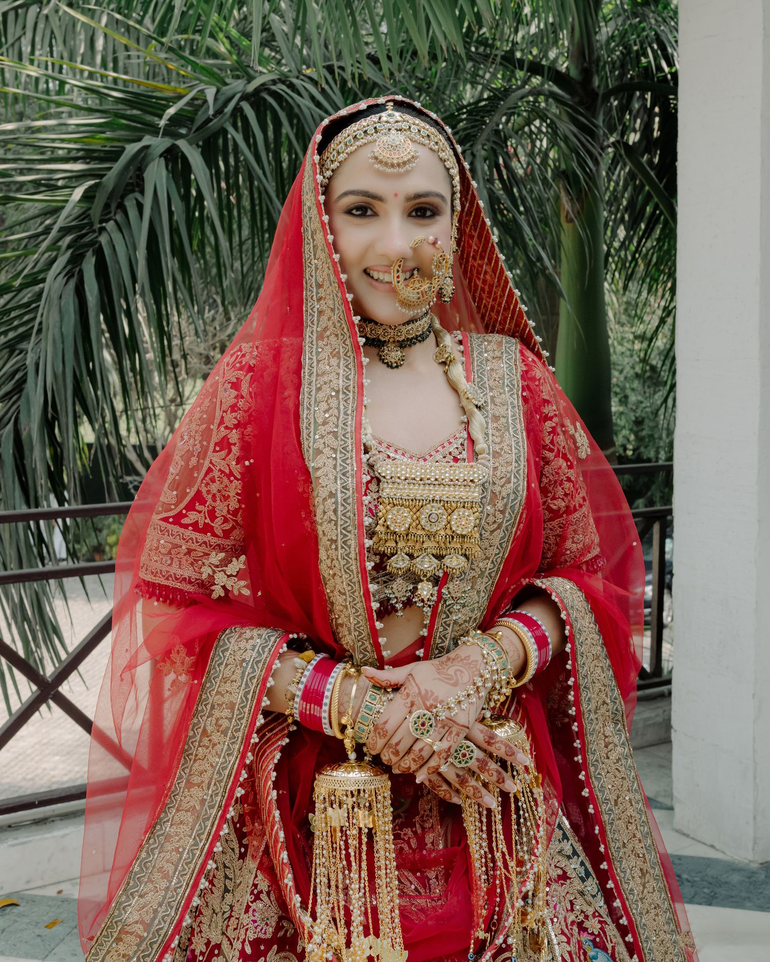 A bride in a red and gold embroidered lehenga with traditional jewelry, dupatta, and mehndi poses outdoors.