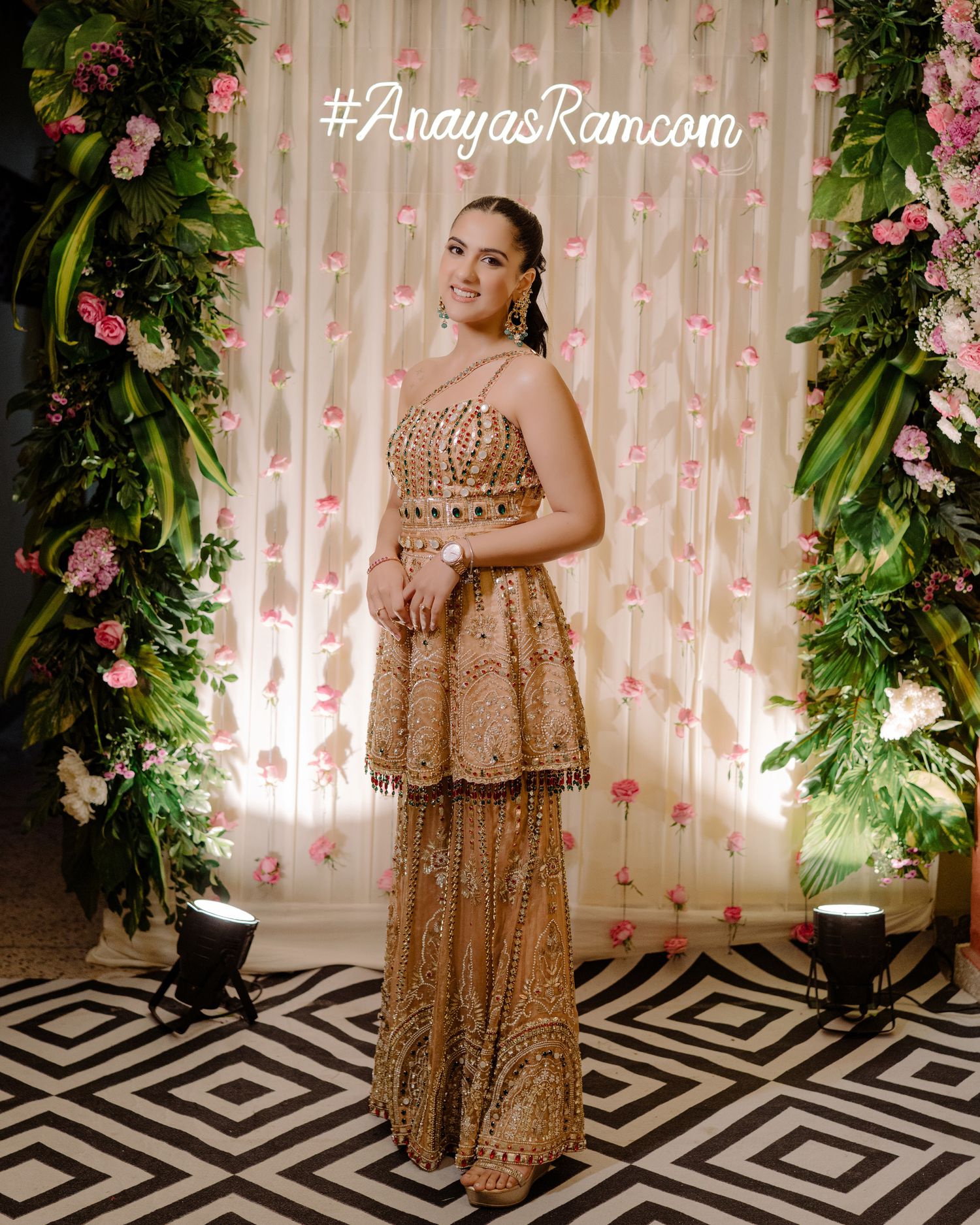 Woman in gold embroidered lehenga poses before floral backdrop at AnayasRam.com event.