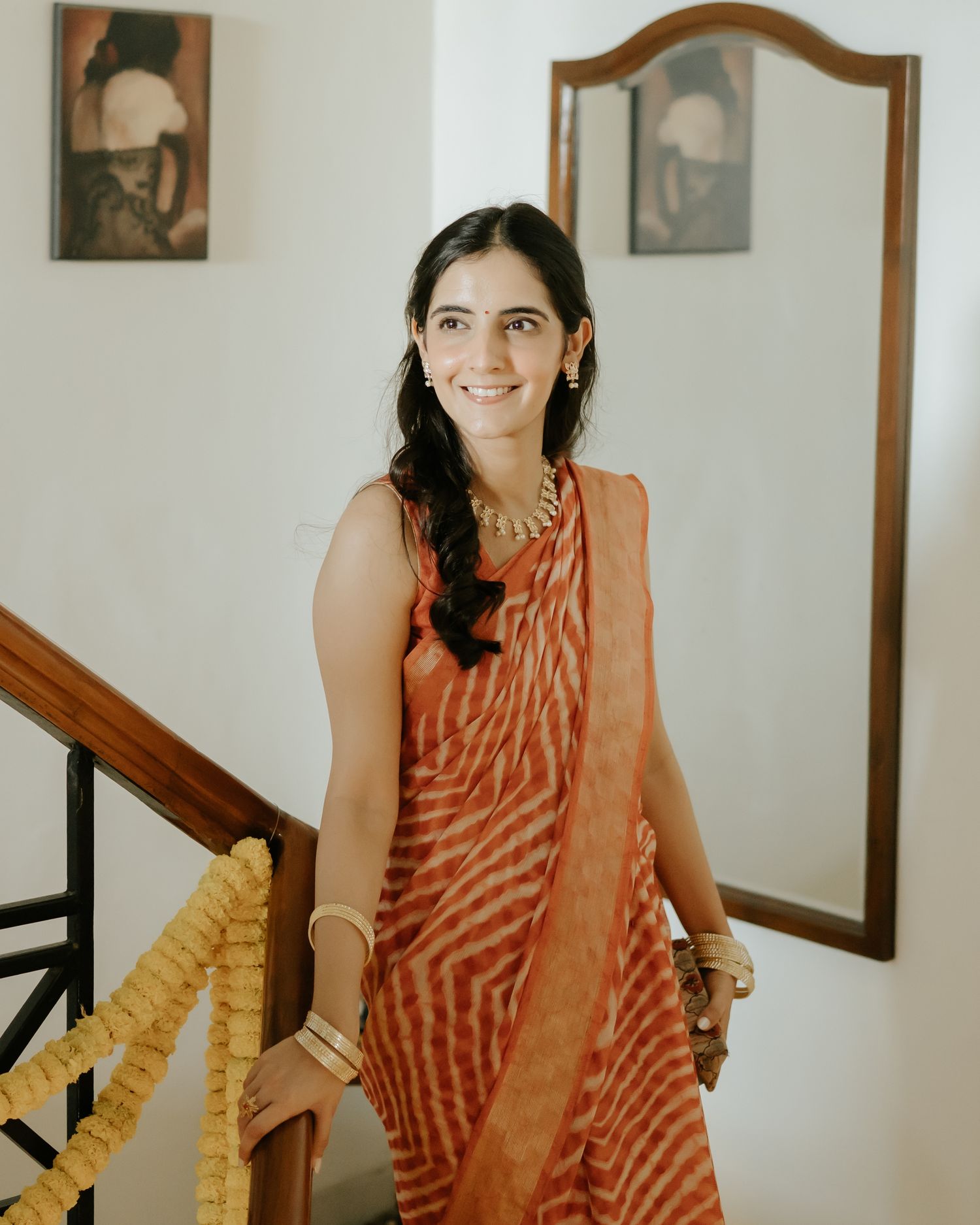 Woman in orange printed saree smiling on staircase with traditional jewelry and artwork on wall behind her.