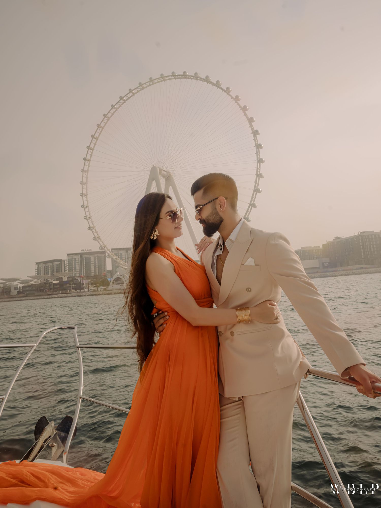 Couple in orange dress and beige suit posing romantically on a yacht with Ain Dubai Ferris wheel backdrop.