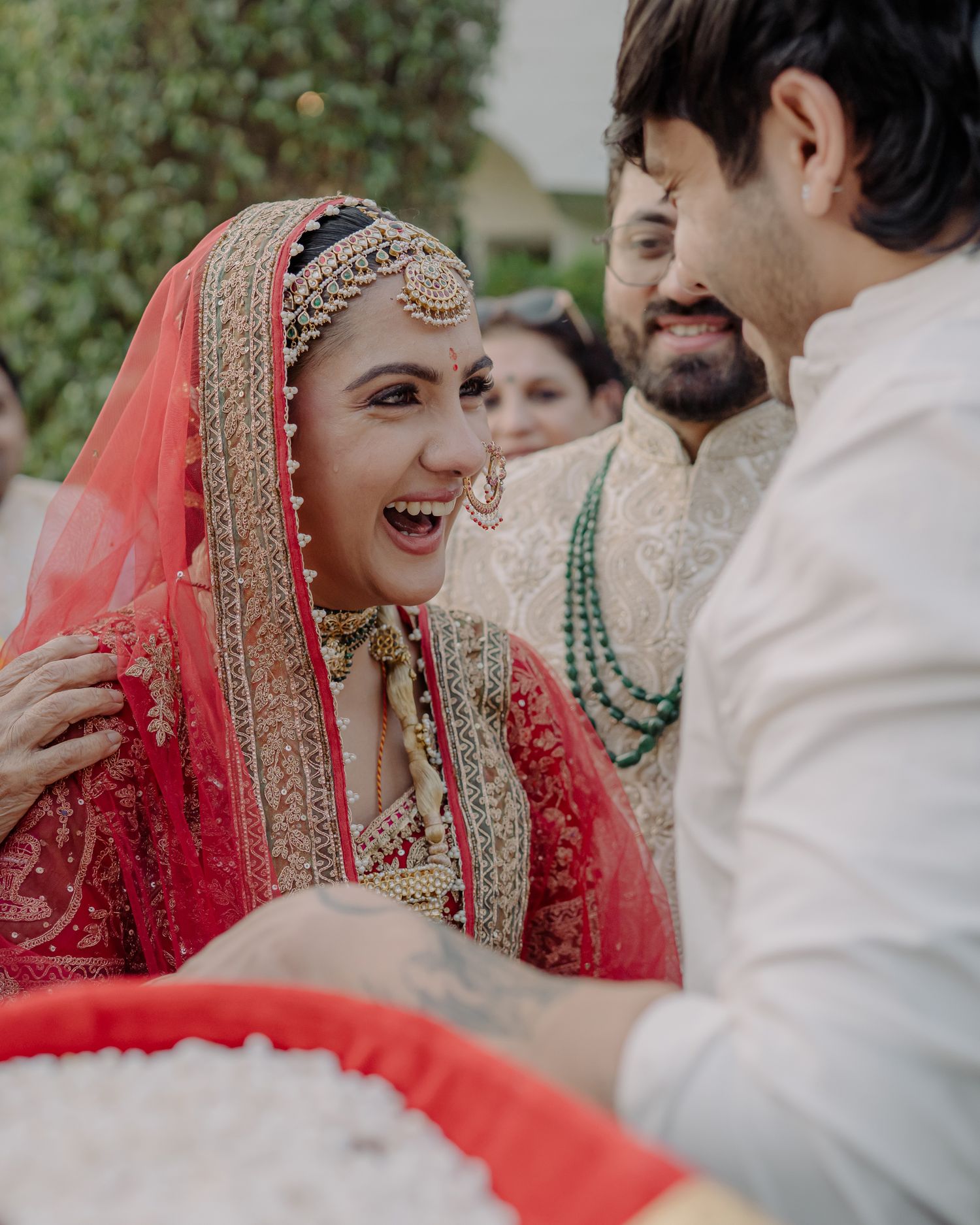 Joyful bride in red bridal lehenga laughing with groom during traditional Indian wedding ceremony.