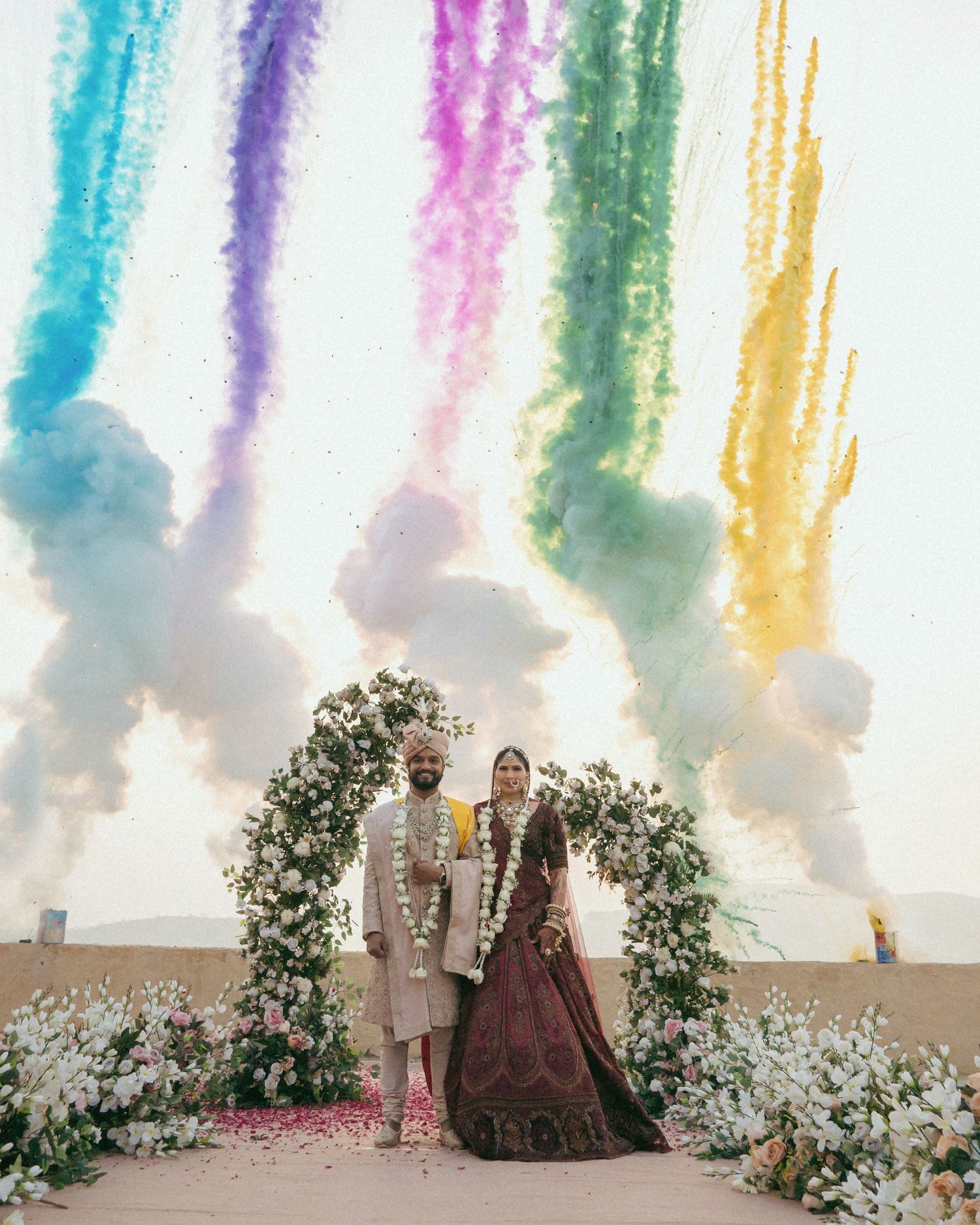 Indian couple in traditional wedding attire stand under colorful rainbow smoke fireworks beside a floral arch outdoors.