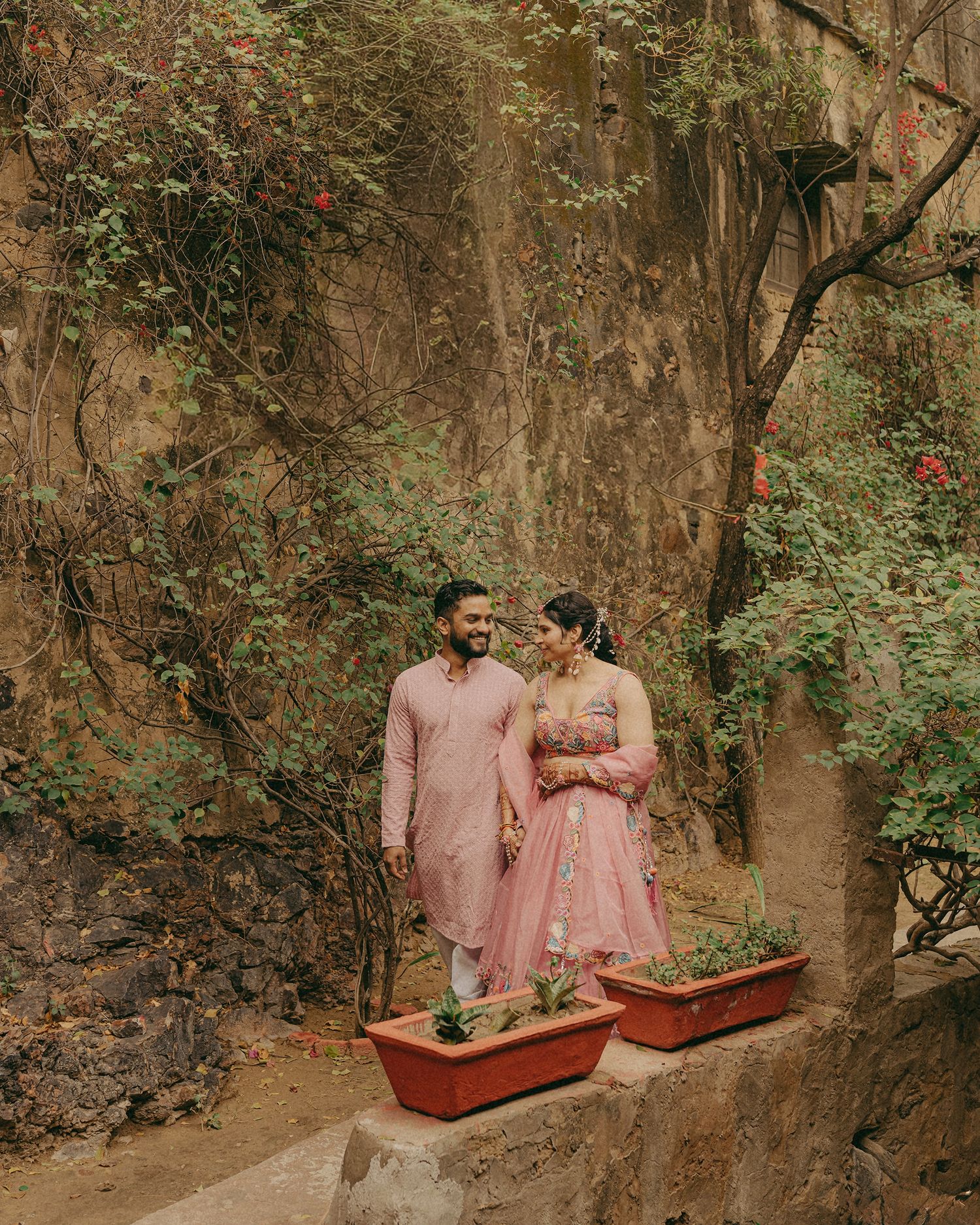 Couple in matching pink Indian attire posing together on stone steps surrounded by lush greenery and climbing vines.