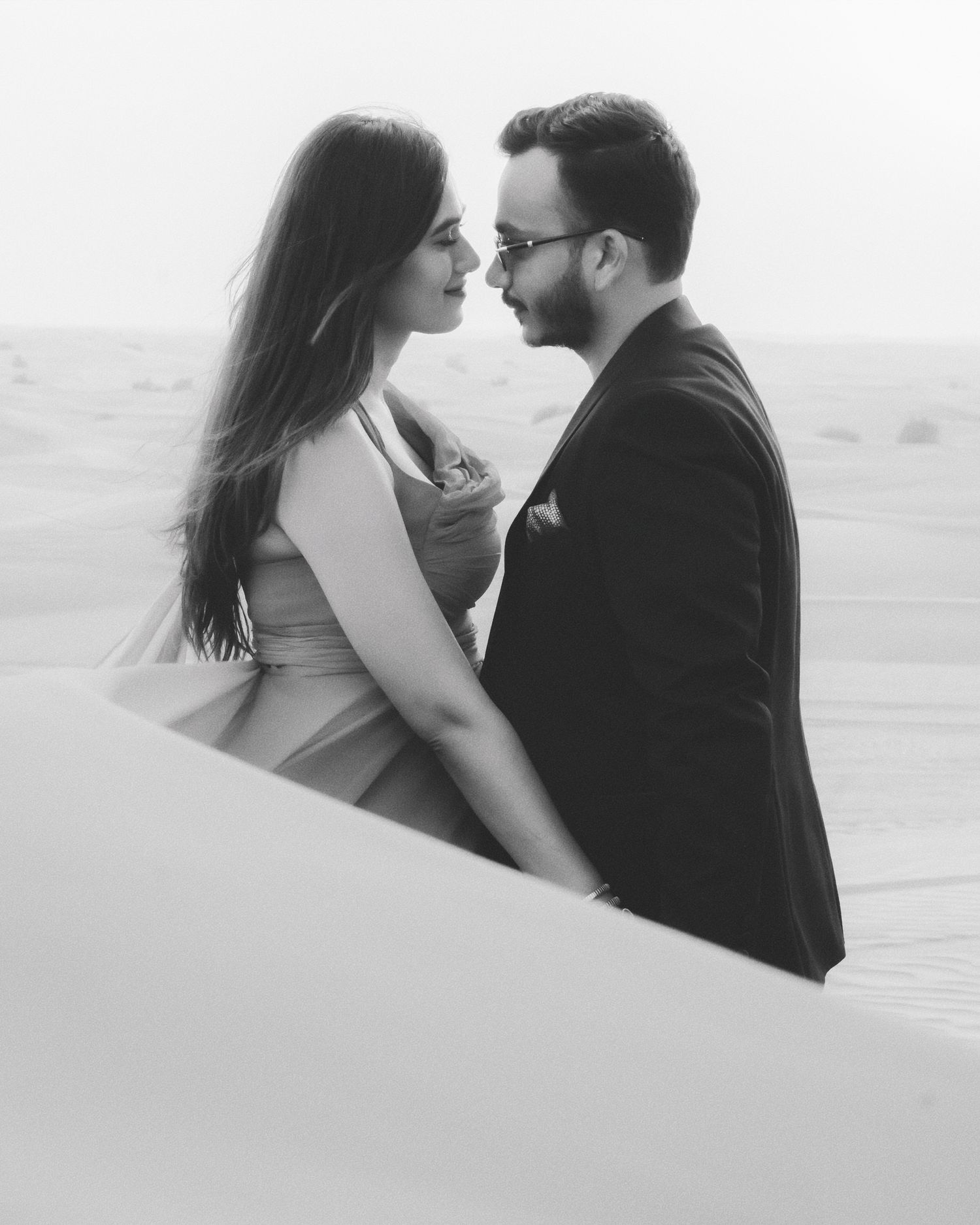 Black and white photo of a couple in formal wear sharing a romantic moment in a desert landscape.