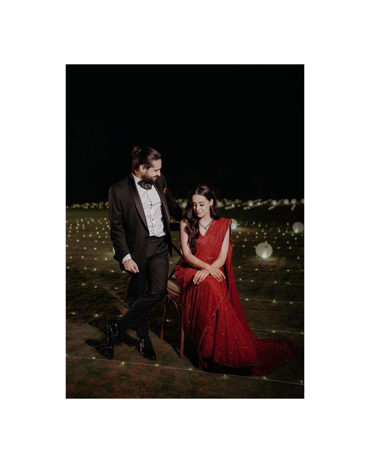 A couple in formal attire share a romantic moment outdoors at night, surrounded by twinkling lights in a dark setting.