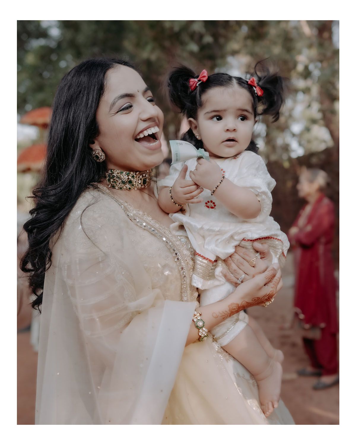Two people in luxurious beige Indian attire share a joyful candid moment outdoors with elaborate jewelry and traditional styling.