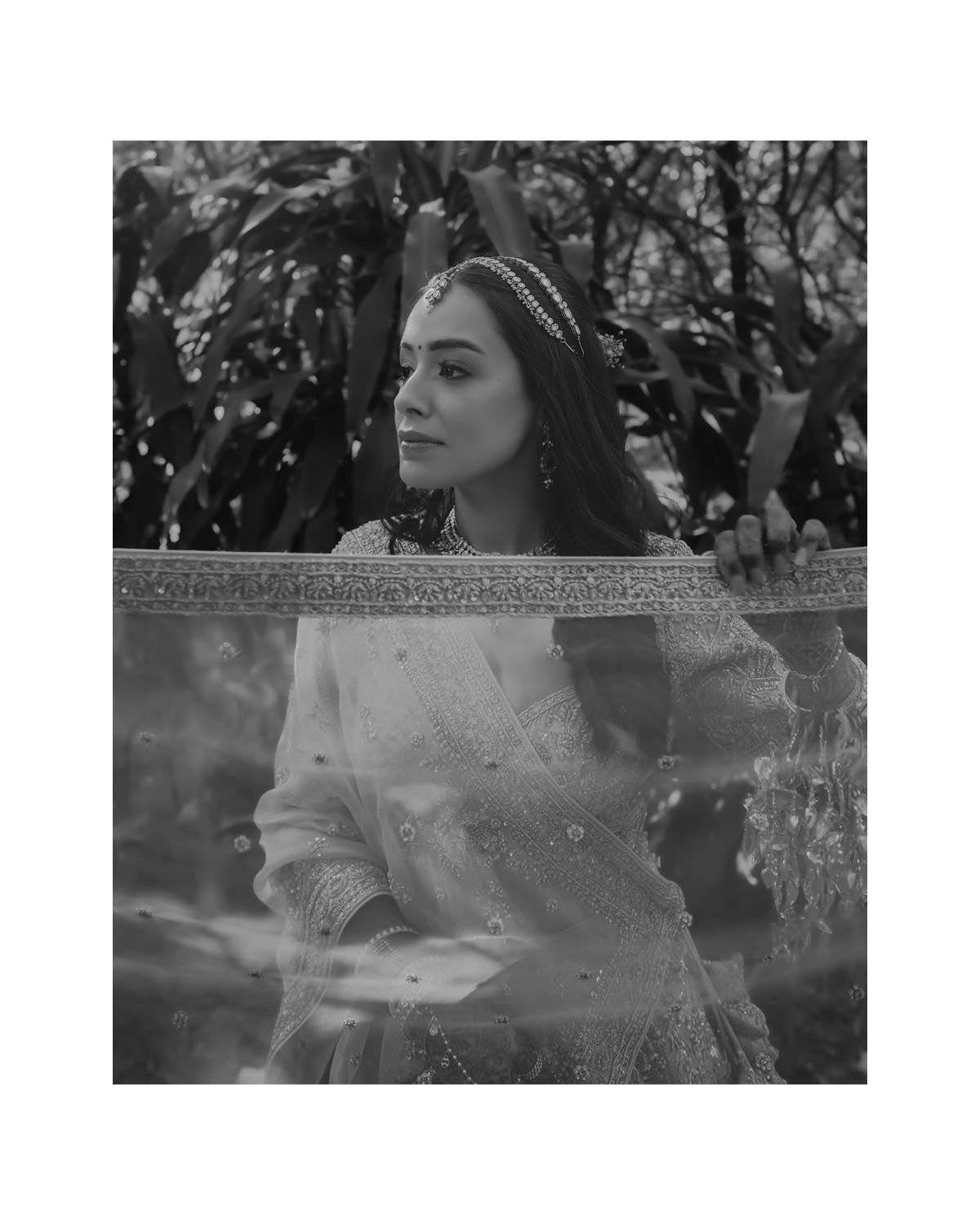 Artistic black and white portrait photo of someone wearing a headband and ornate dress against a leafy background.