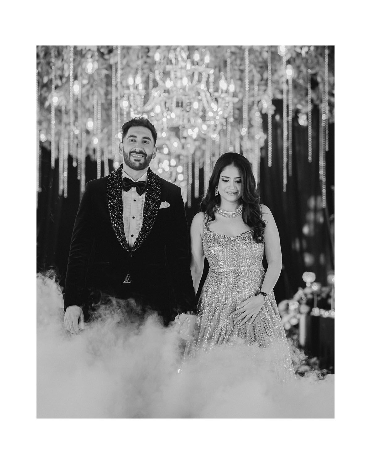 Black and white portrait of couple standing together amid hanging crystal decorations and fog effects.