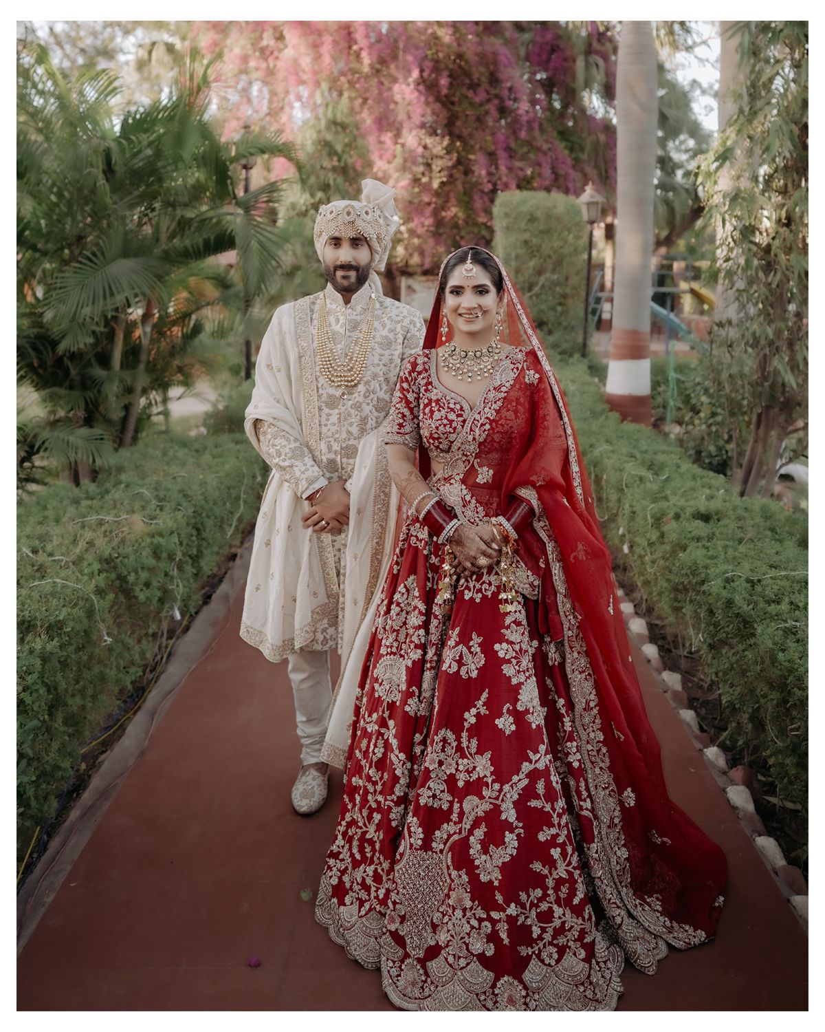 A full-length shot of a wedding couple in traditional Indian attire standing on a garden pathway.