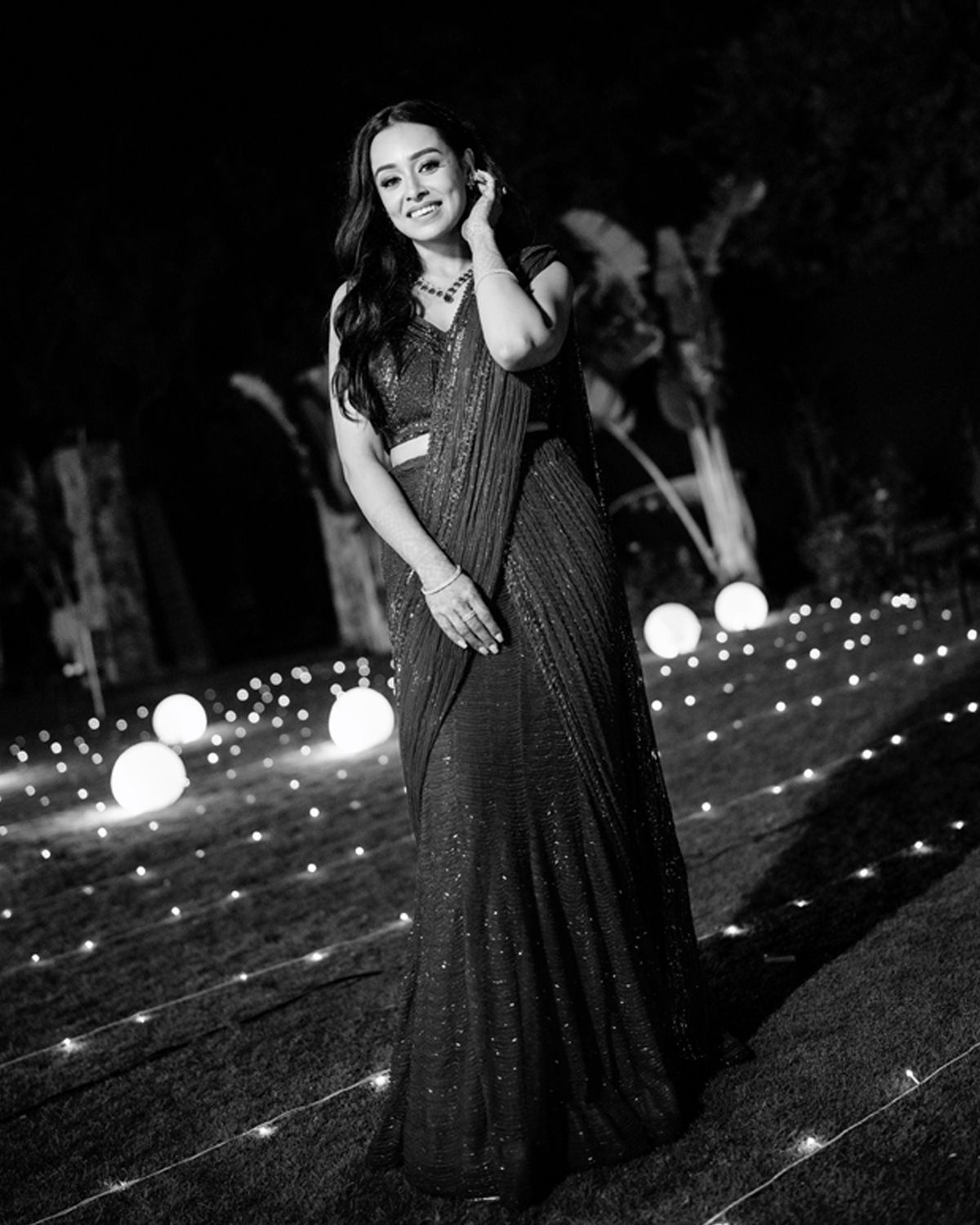 A woman in an elegant black sari poses at night with glowing lights and decorative elements in the background.