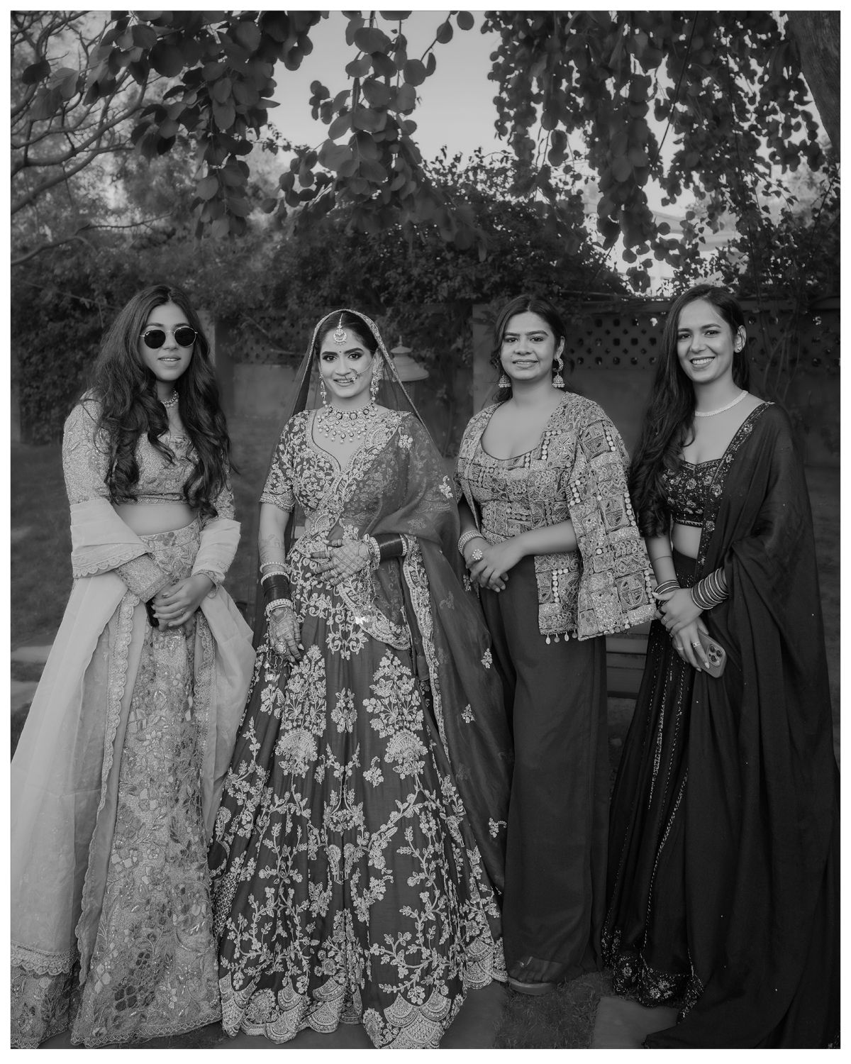 Four women dressed in traditional Indian formal wear pose together in a garden setting.