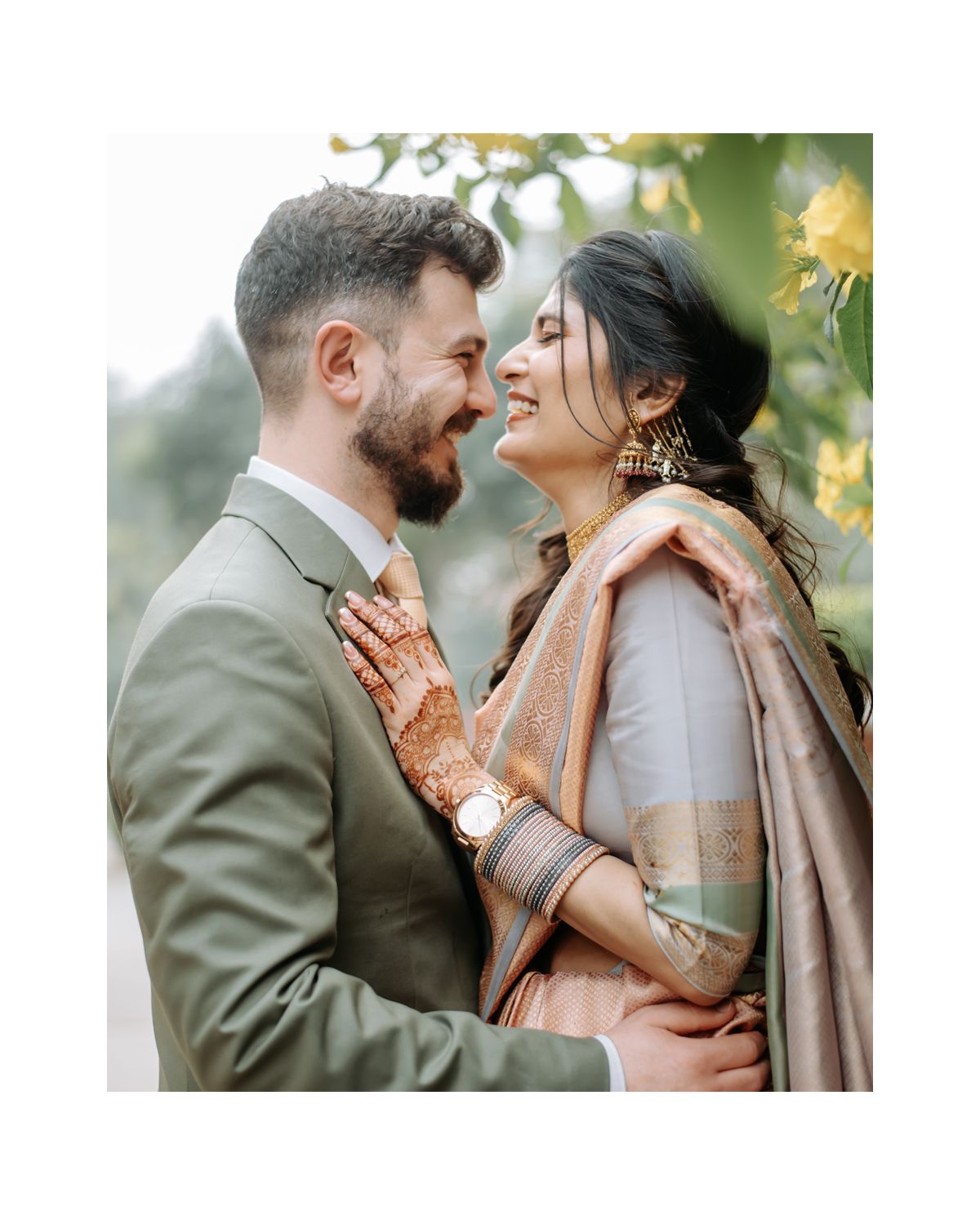 Intimate portrait of couple in traditional South Asian wedding attire against green foliage.