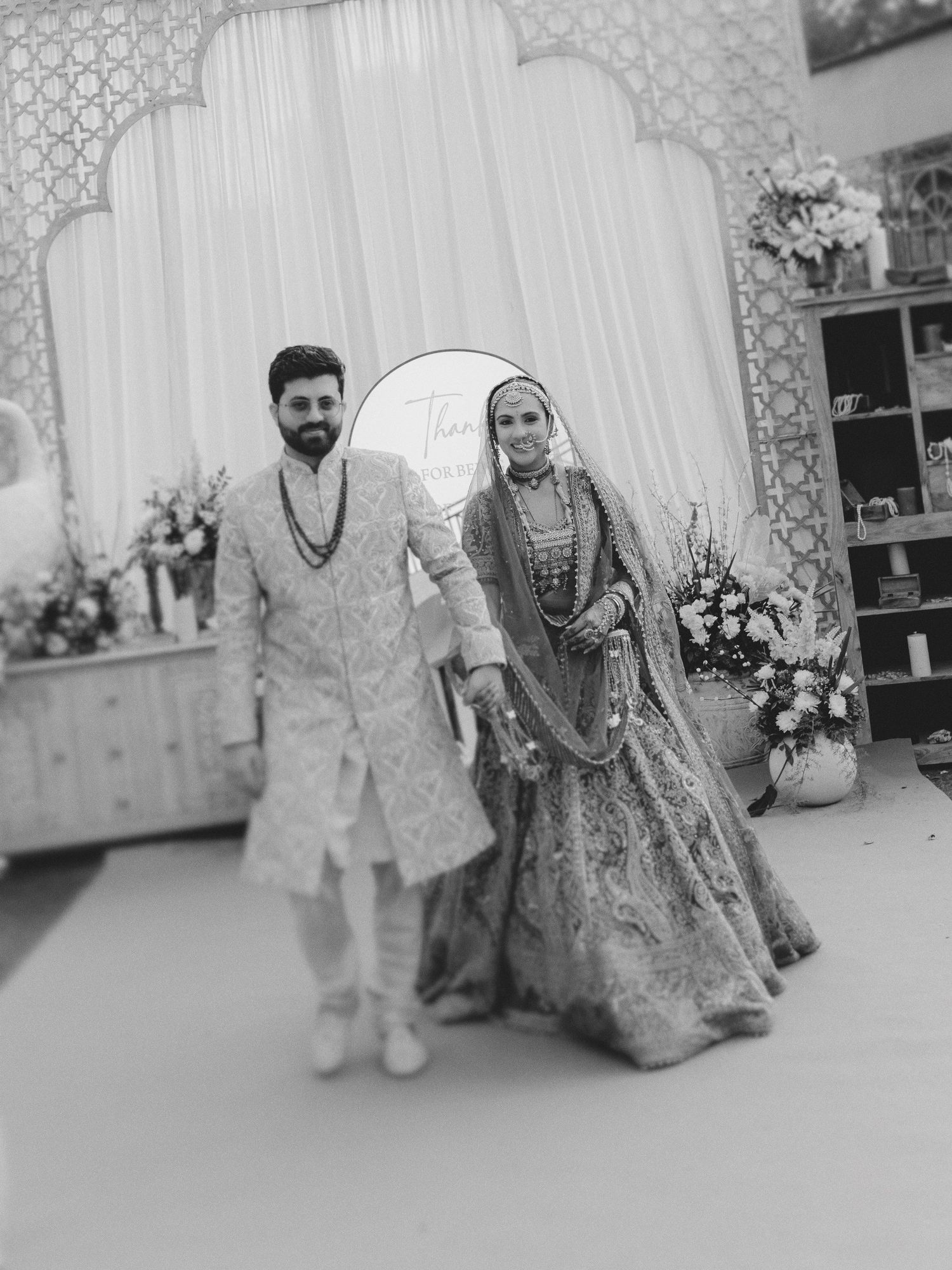 Black and white photo of an Indian bride and groom in traditional wedding attire posing together at an elegant venue.