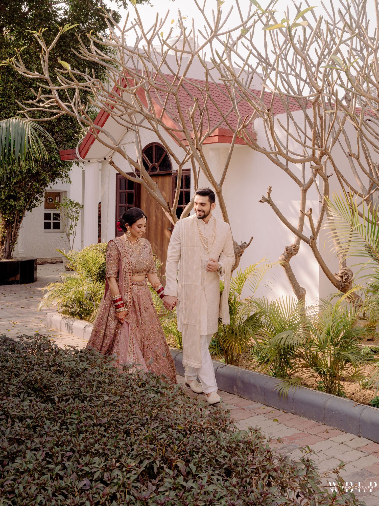Indian bride and groom in traditional attire walking hand in hand in a lush garden outside a white colonial venue.