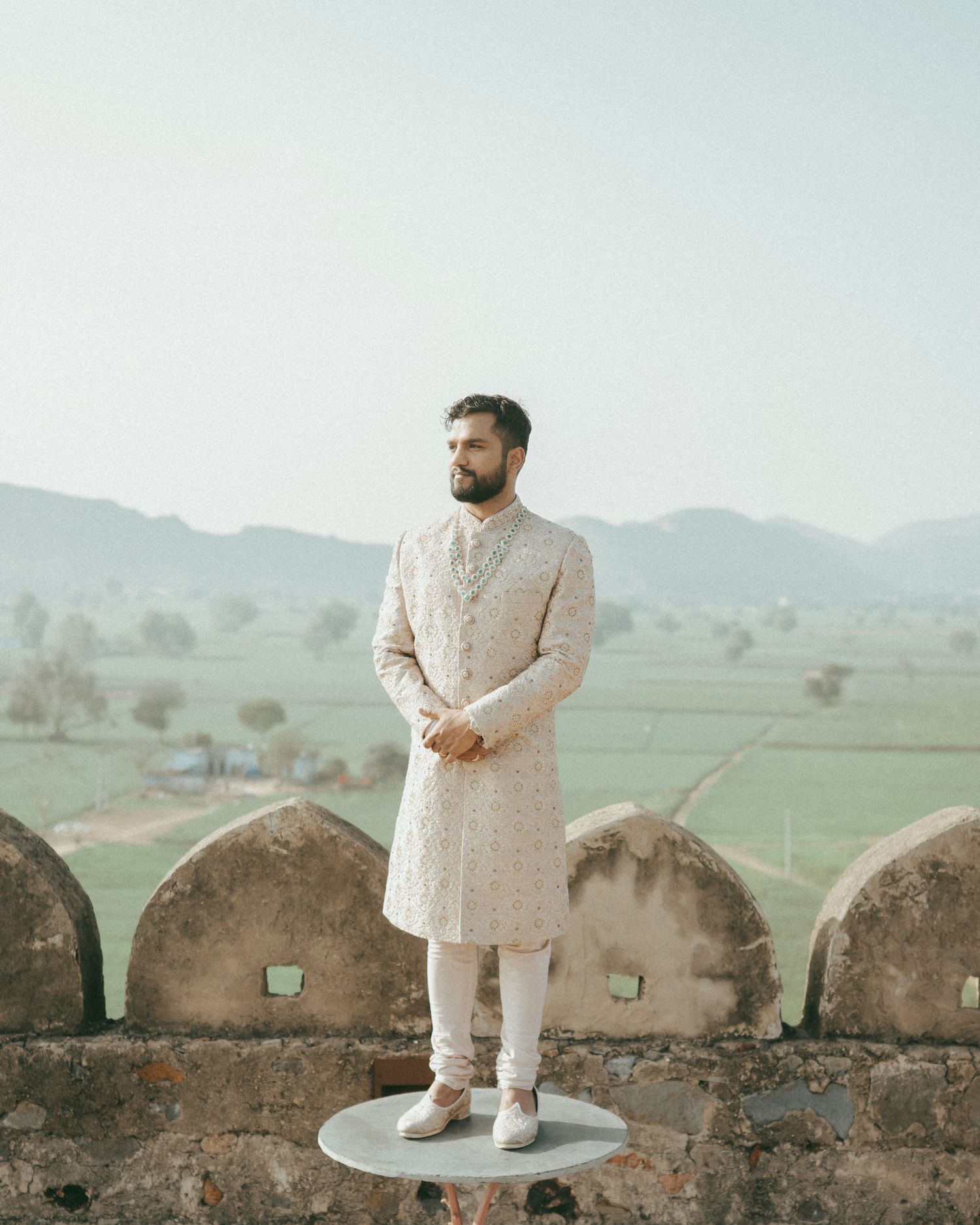 Man in cream embroidered sherwani stands on stone fort wall with green landscape and misty mountains behind.