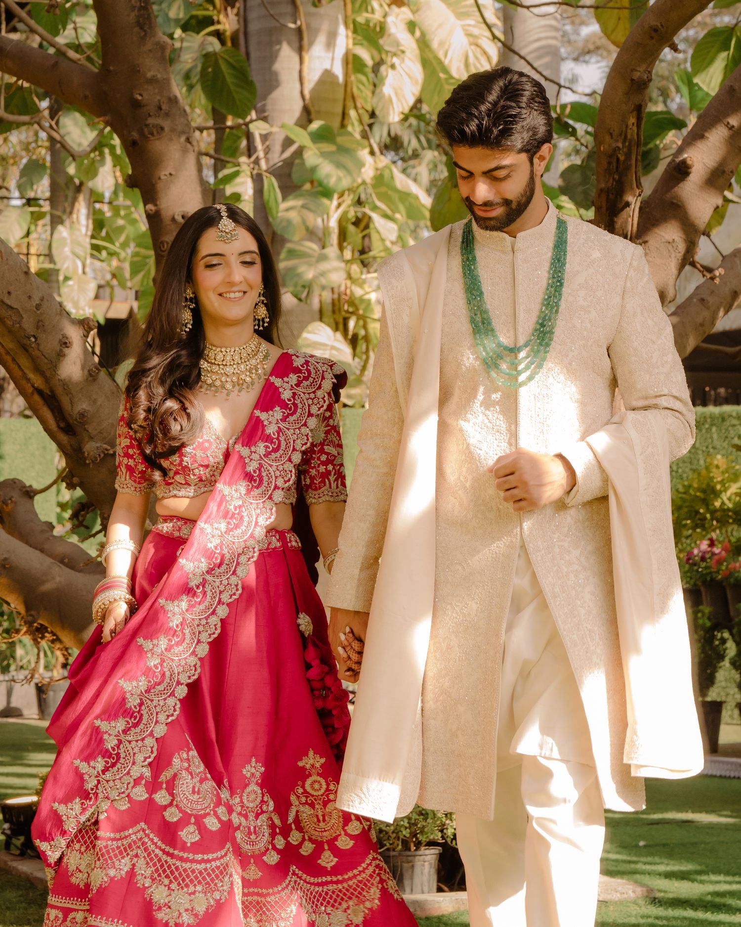 Couple in traditional Indian wedding attire, bride in red lehenga with gold embroidery, groom in cream sherwani with green mala.