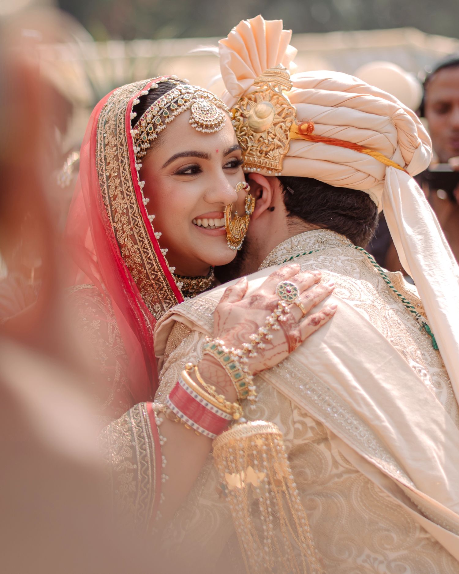 A smiling Indian bride in gold jewelry and pink dupatta embraces groom in white sherwani and turban at wedding.