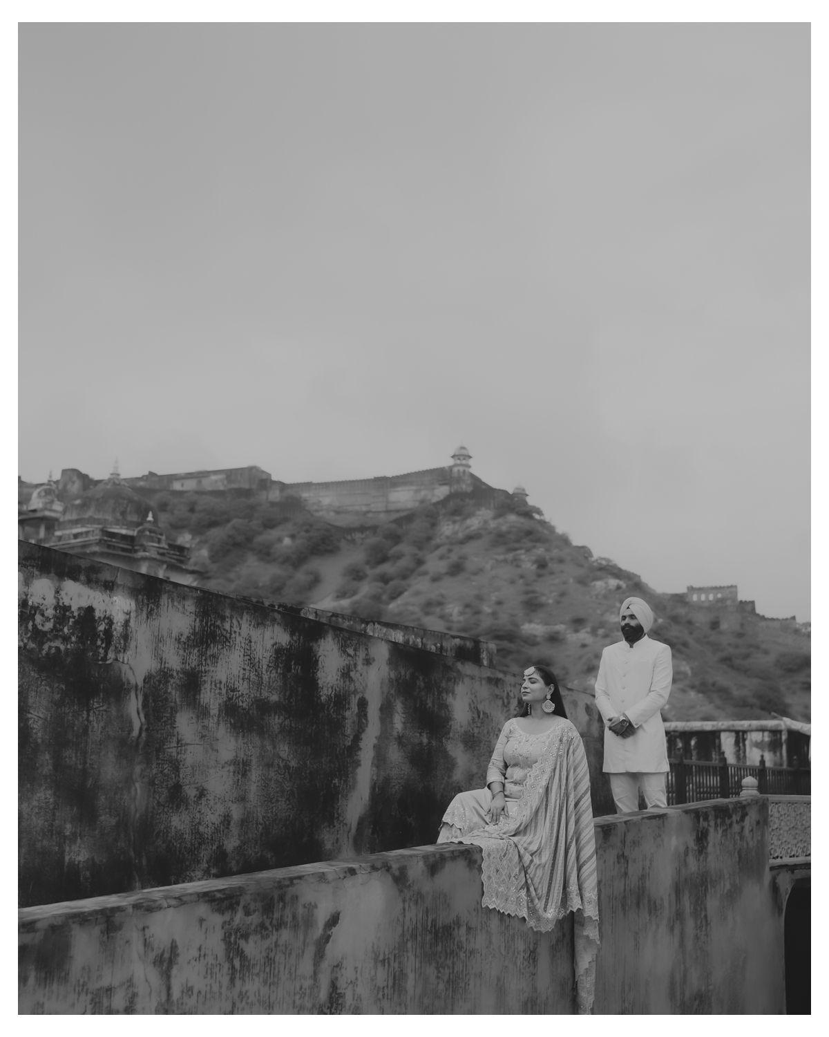 Artistic black and white photo of a couple posing on ancient fort walls with mountains in background.