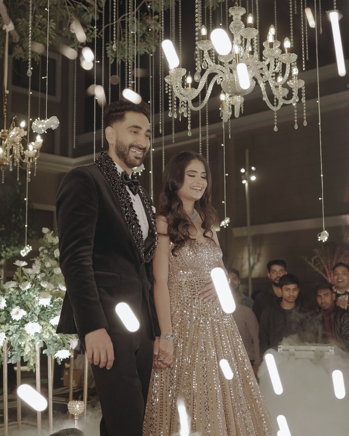 Glamorous celebration scene with sparkling gown and tuxedo under hanging crystal chandeliers and floral arrangements.