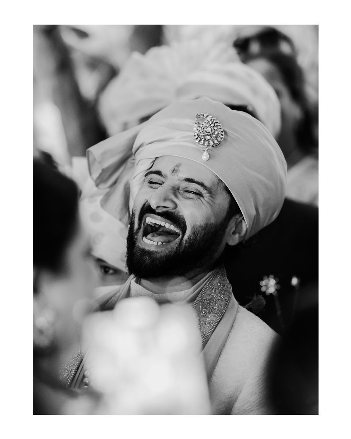 A person in traditional wedding attire laughs joyfully while wearing a white turban adorned with an ornate brooch in black and white.